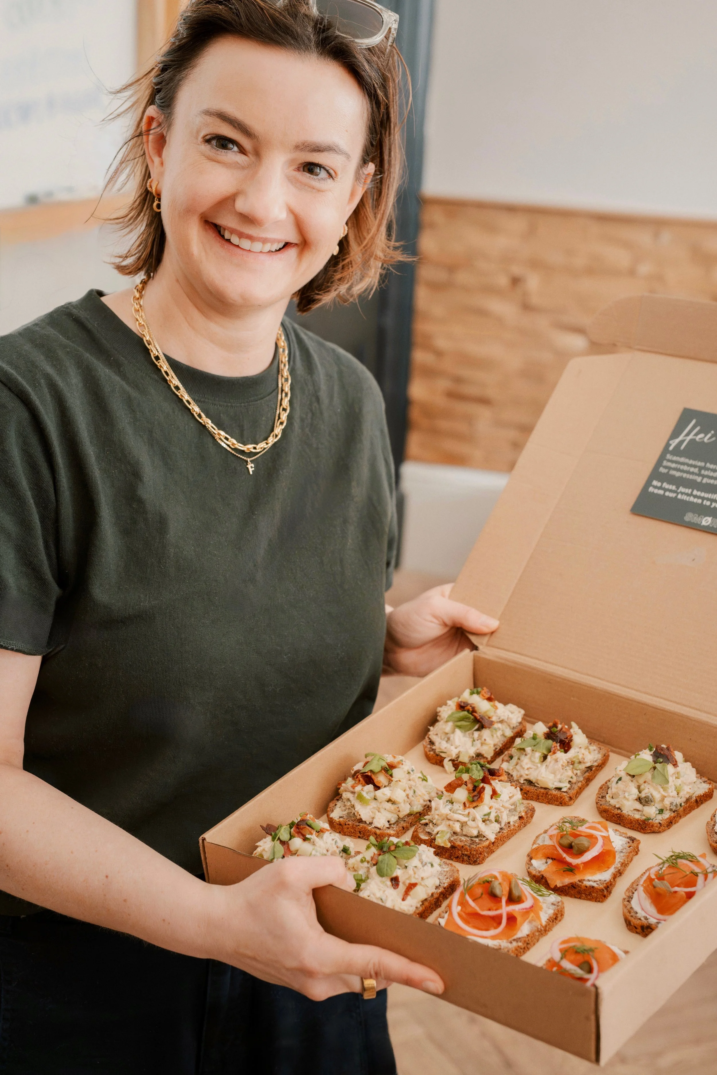 A woman with short brown hair, wearing a black T-shirt and gold jewelry, holds a pizza box filled with small, decorated pizzas topped with various herbs and ingredients.