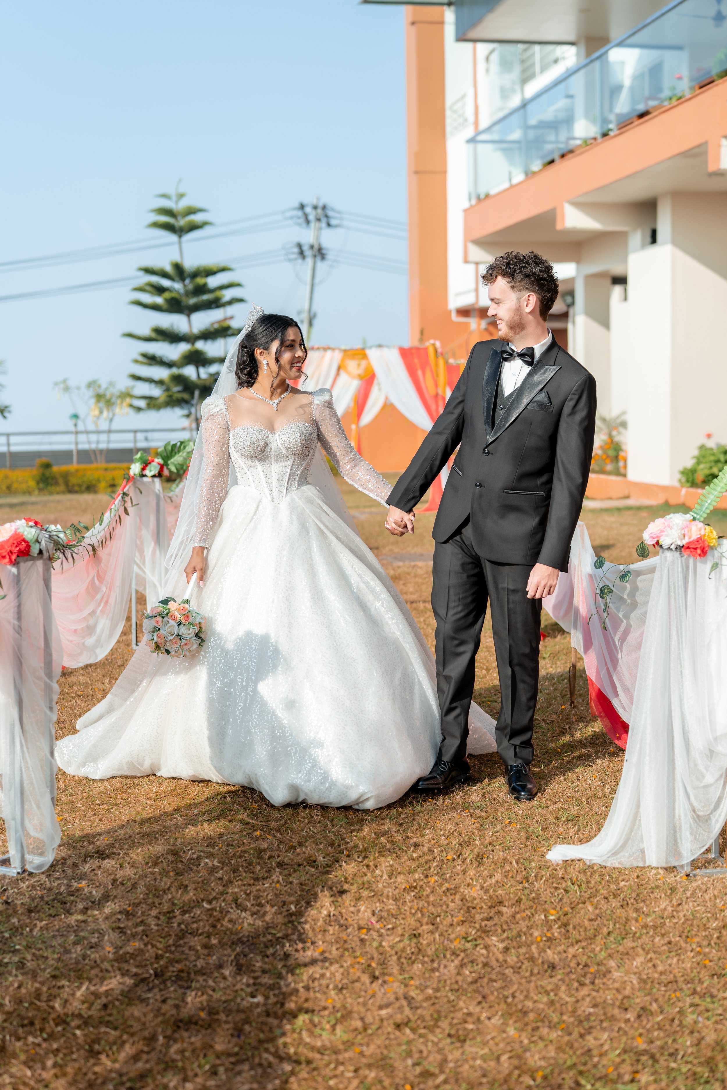 A bride in a white wedding gown and a groom in a black tuxedo holding hands outdoors during their wedding ceremony, with decorative flowers and fabric draping visible.