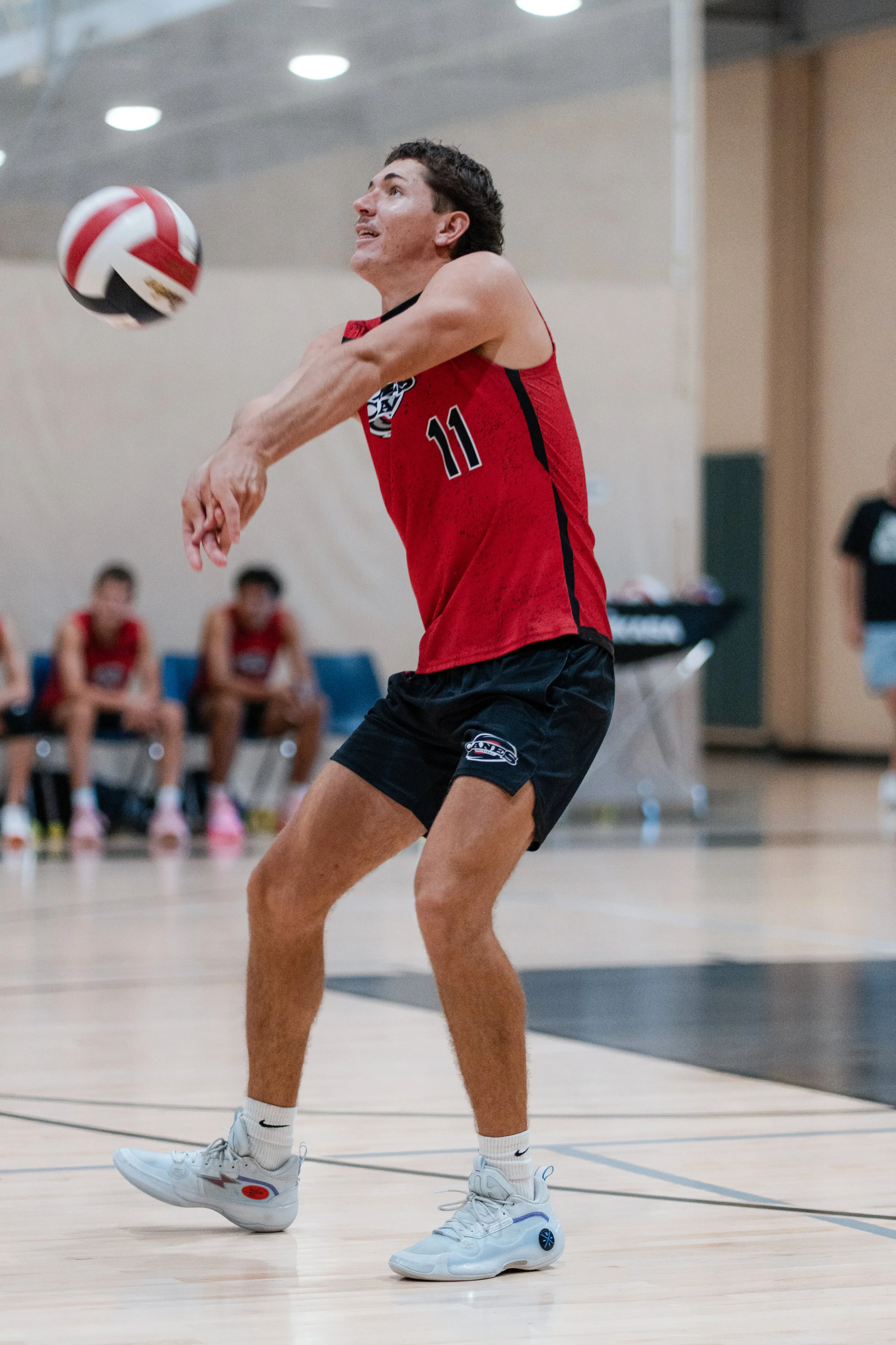 A male volleyball player in a red jersey with the number 11 is hitting a volleyball indoors, with teammates sitting on a bench in the background.
