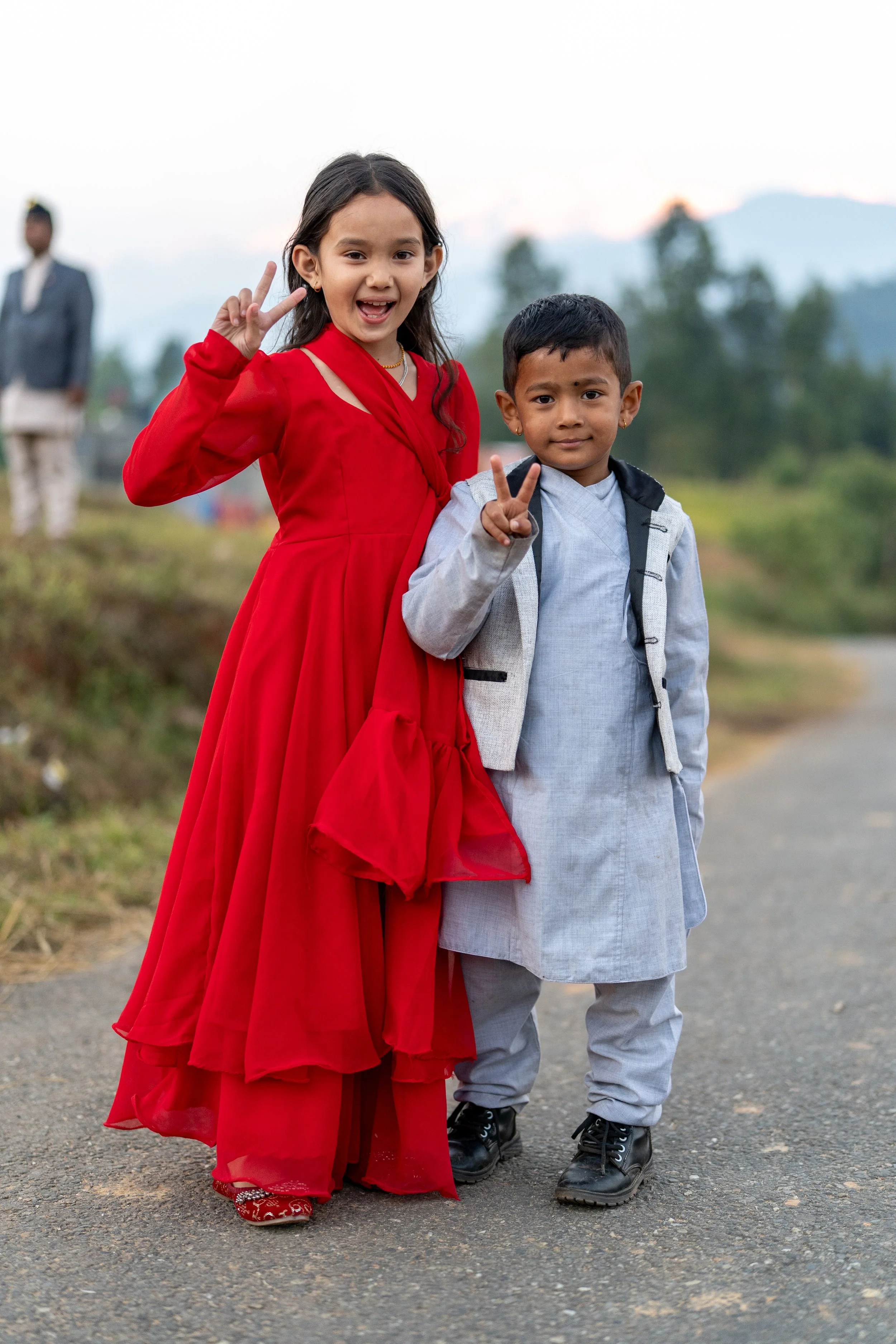 Two children, a girl in a red dress and a boy in traditional attire, standing on a rural road making peace signs with their fingers.