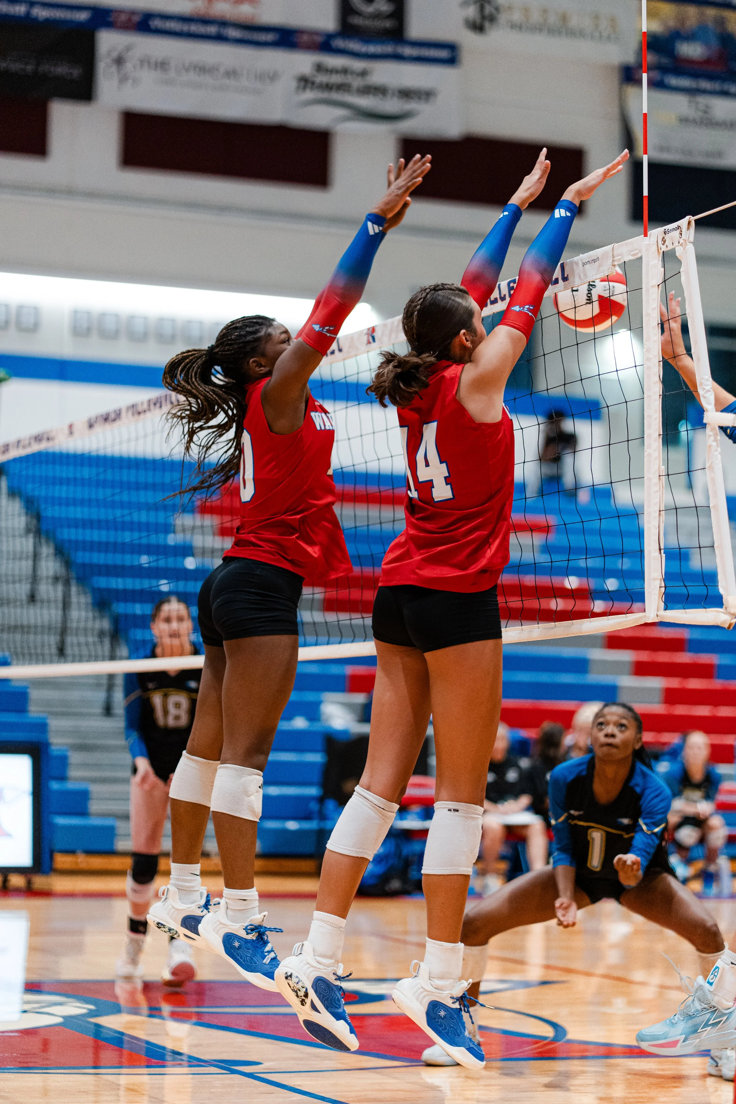 Two female volleyball players in red jerseys jumping to block the ball at the net during a match, with other players and spectators in the background.