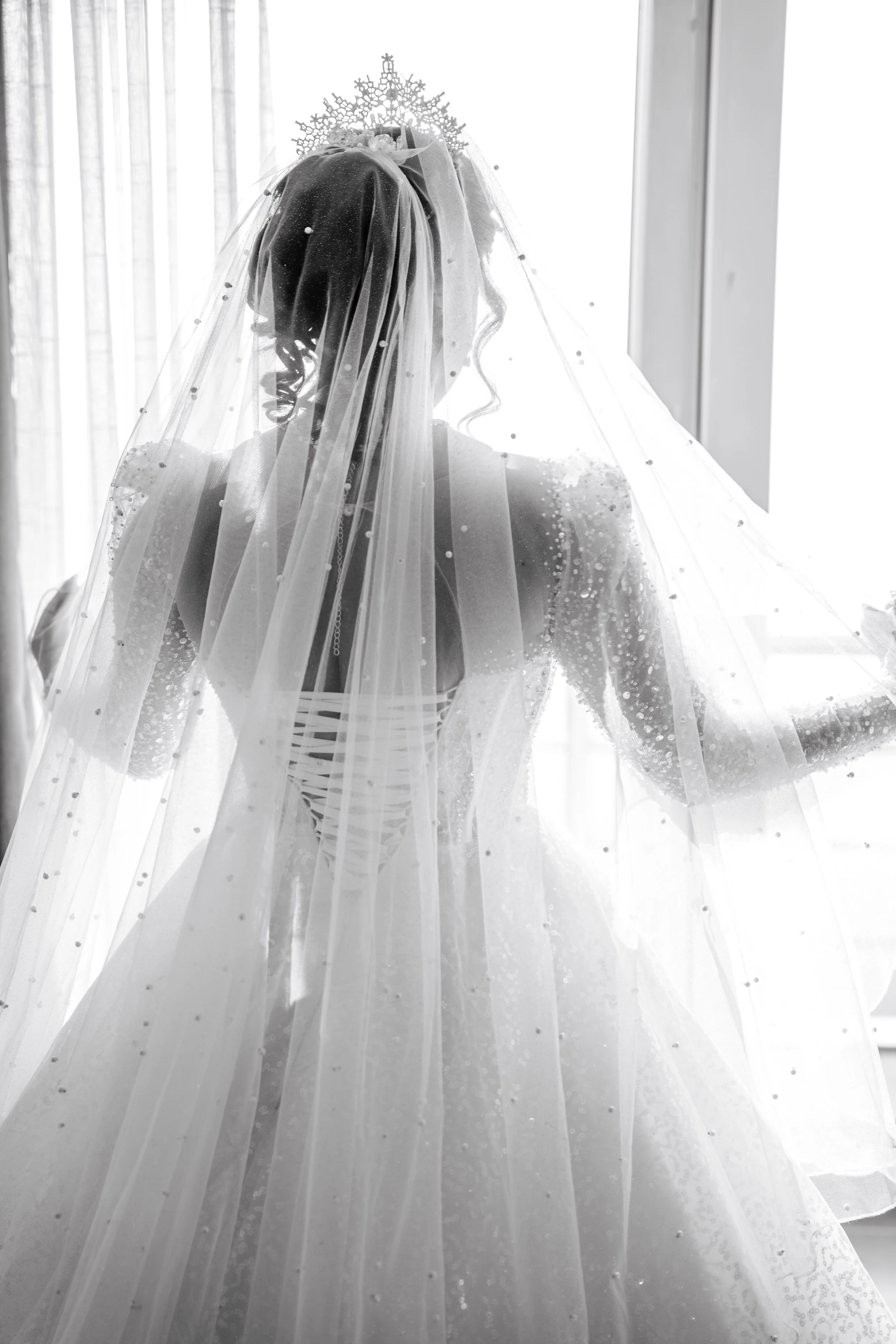 A bride dressed in a wedding gown and veil, wearing a tiara, standing near a window with curtains, back view.