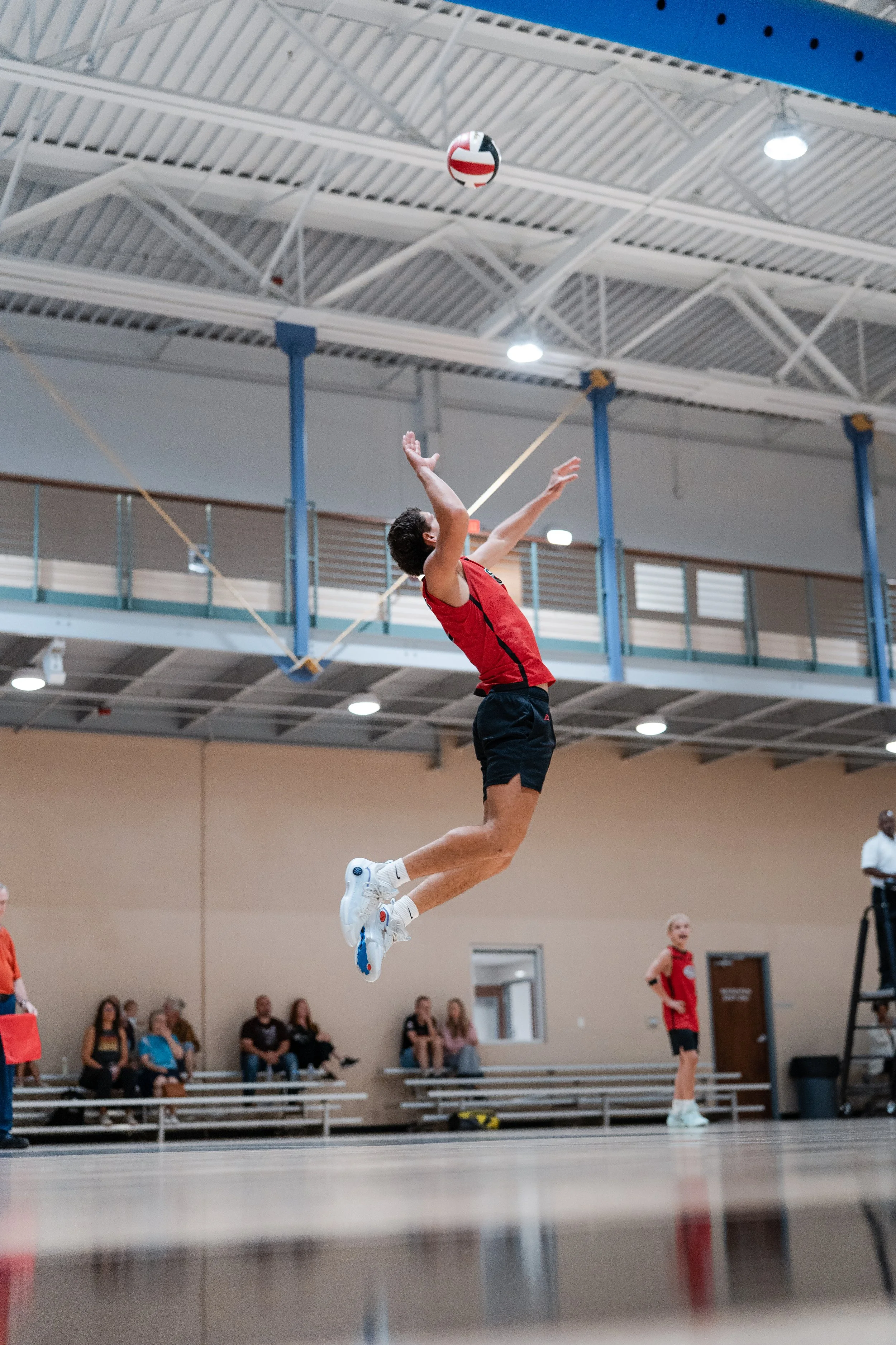 A male volleyball player is jumping to spike the ball in an indoor gym with spectators watching.