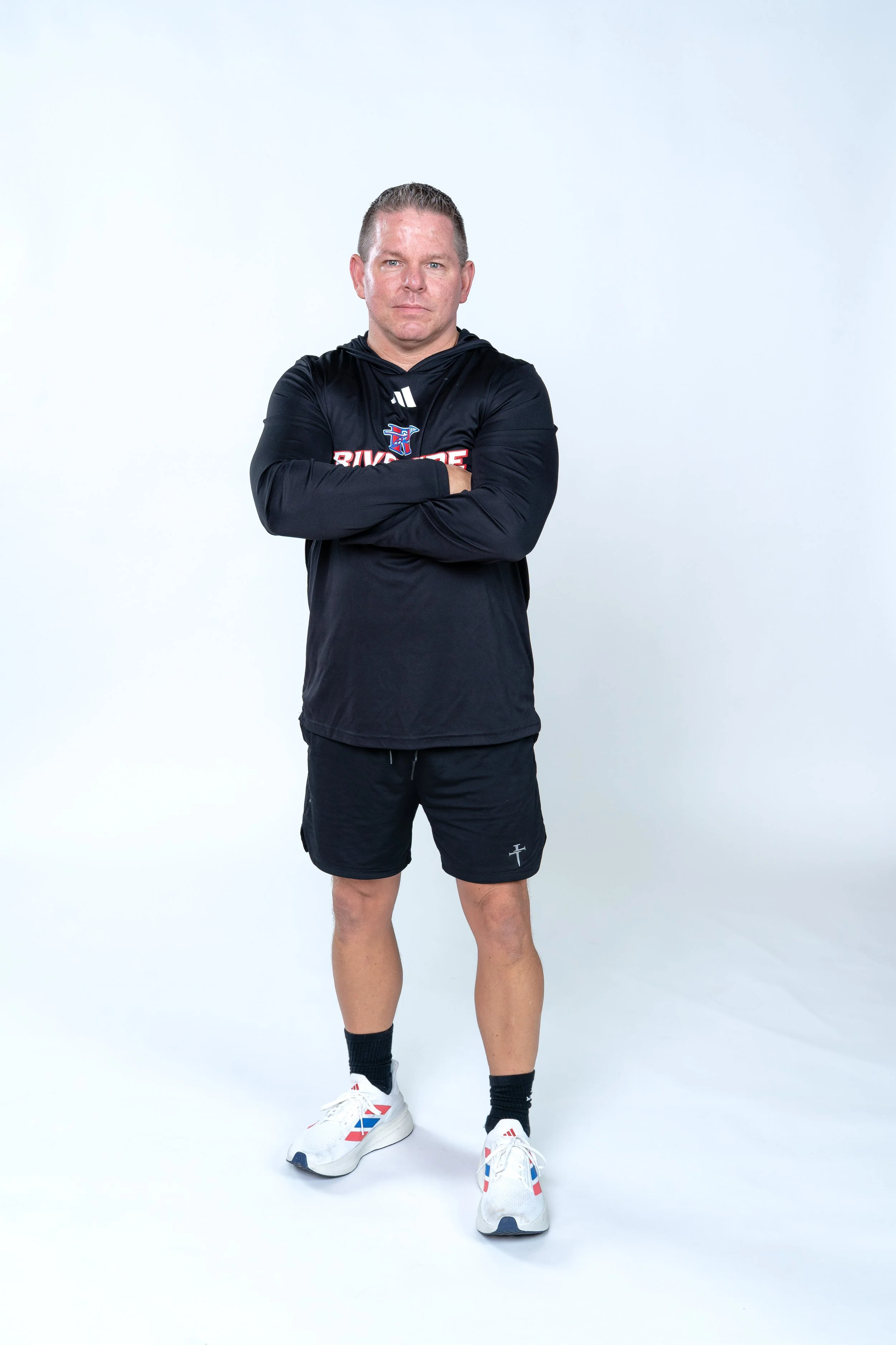 A man standing with arms crossed, wearing a black hoodie, black shorts, black socks, and white athletic shoes, against a plain white background.