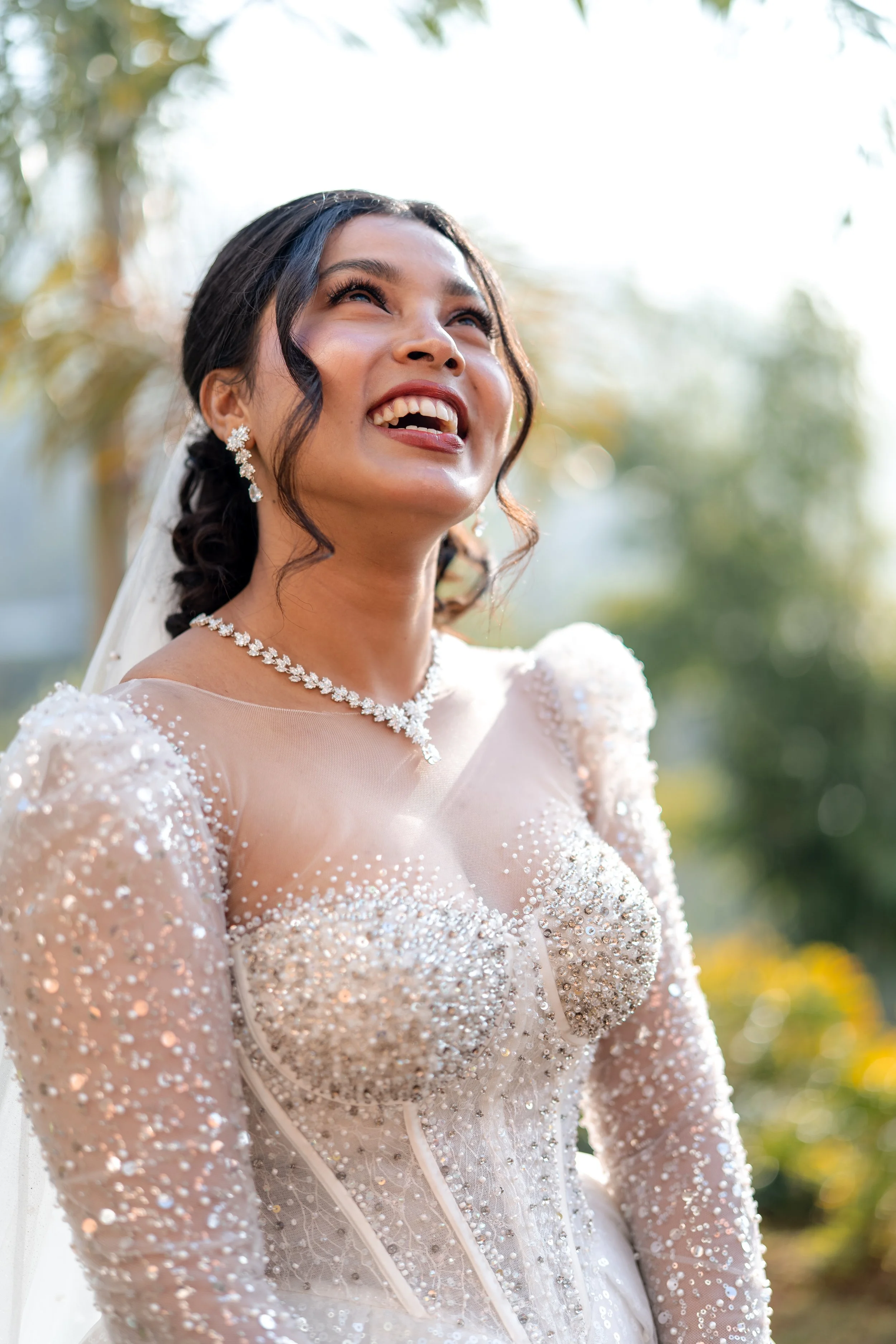 A bride wearing a white, pearl-embellished wedding dress with lace details, a diamond necklace, and earrings, smiling joyfully outdoors with trees and sunlight in the background.