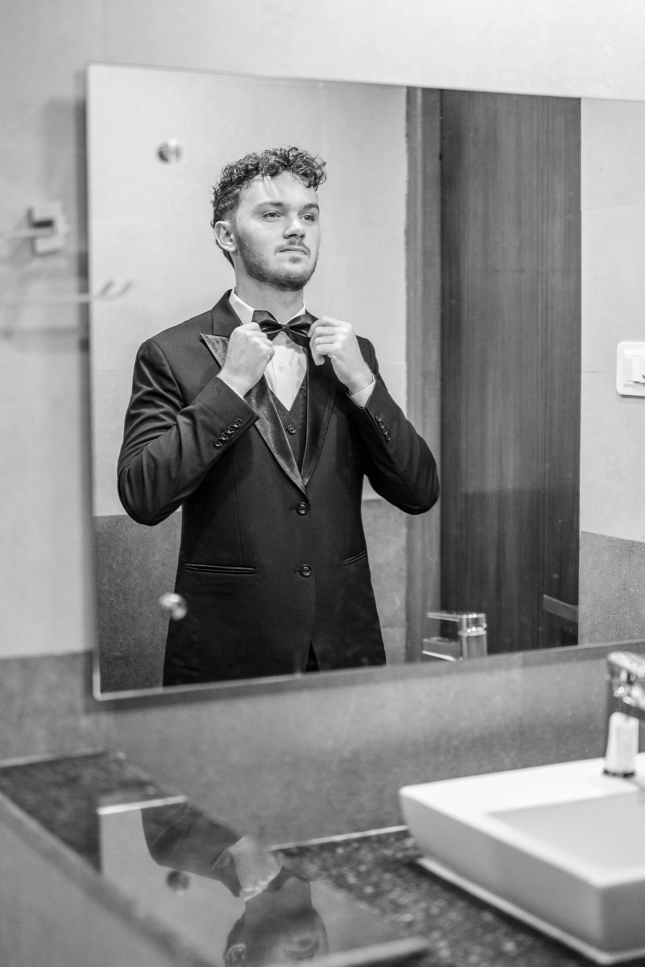 A young man in a tuxedo adjusting his bowtie, looking at himself in a bathroom mirror.