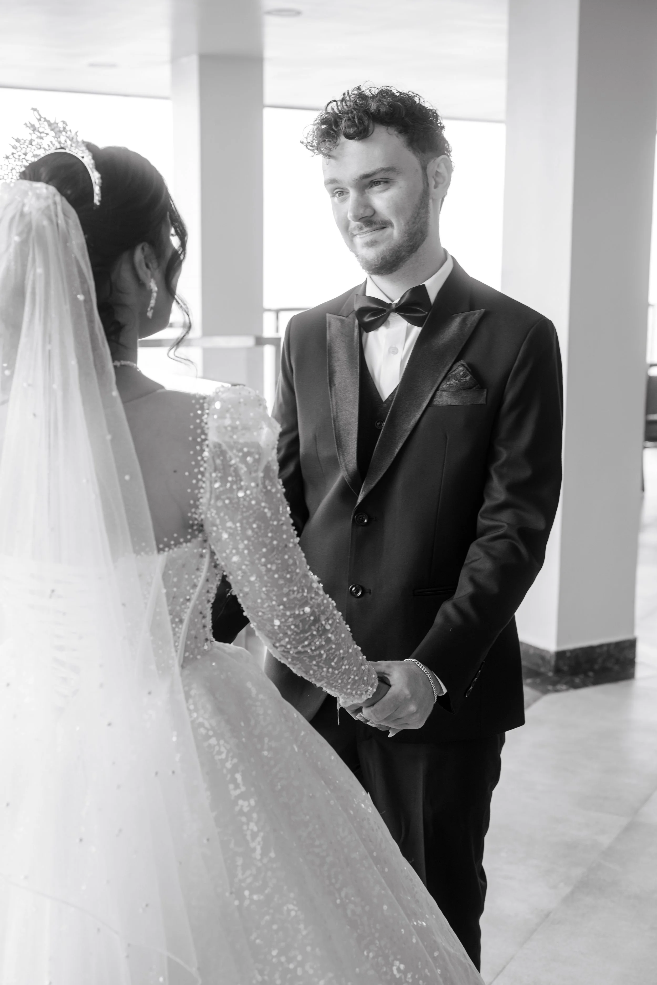 A black and white photo of a bride and groom exchanging vows, holding hands, indoors with modern architecture in the background.