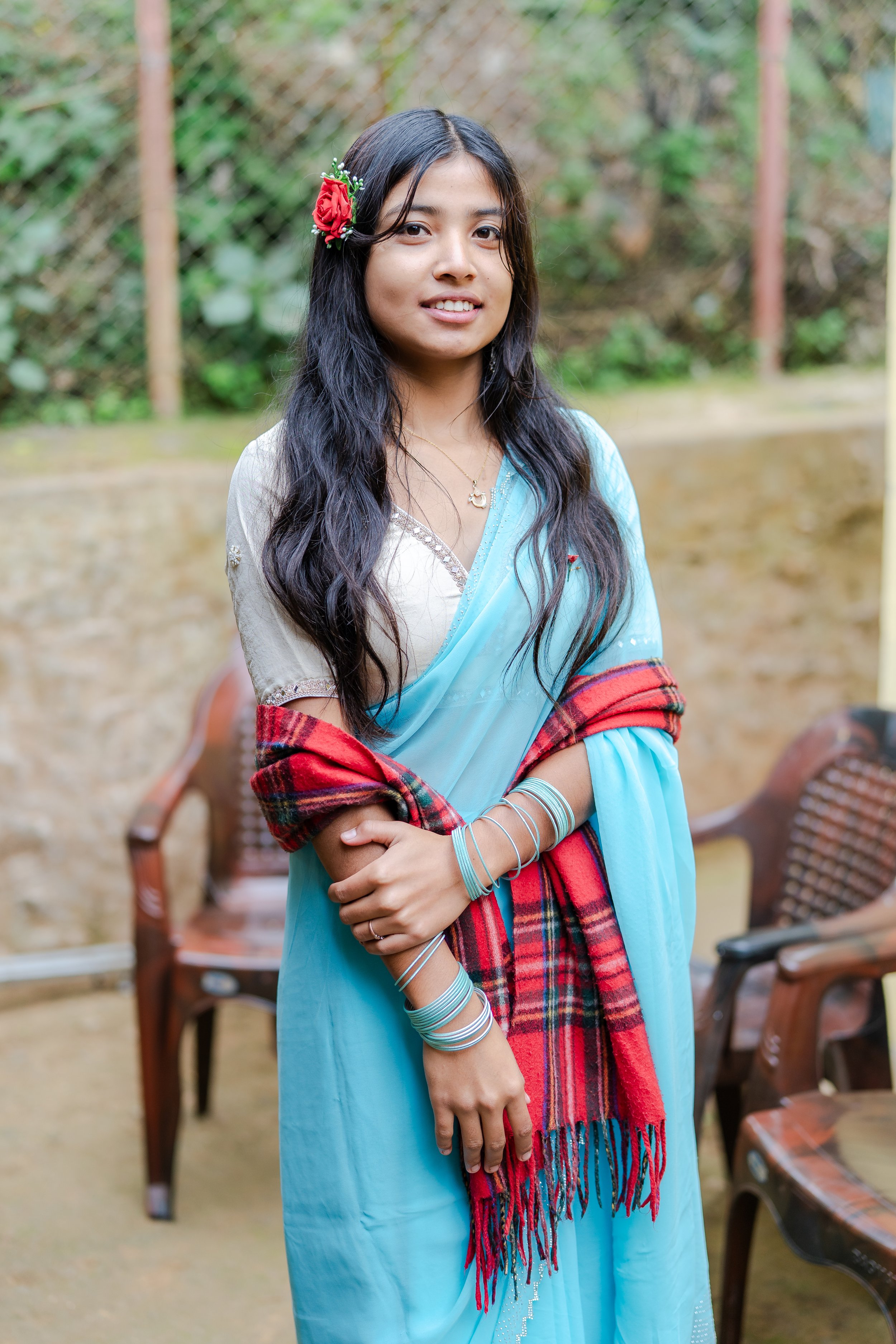 A woman wearing a light blue traditional sari with a red plaid shawl, accessorized with silver bangles, a necklace, and red flower hairpiece, standing outdoors with chairs and greenery in the background.