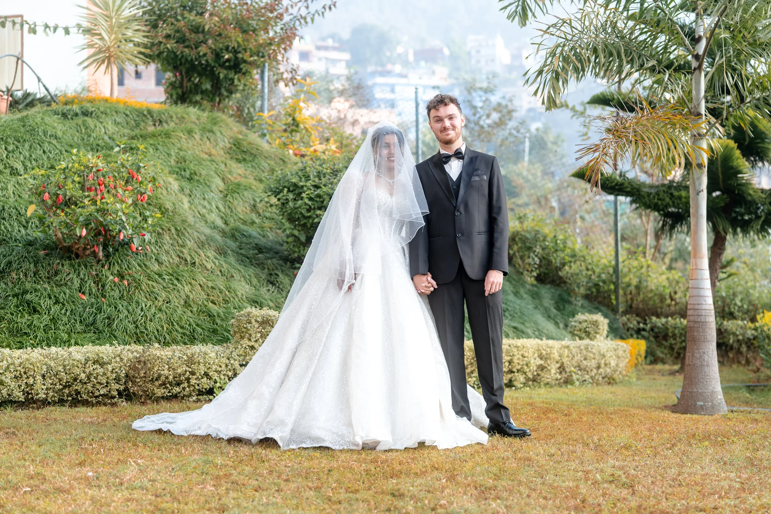 Bride and groom standing outdoors on a grassy lawn, holding hands, with trees and shrubs in the background.