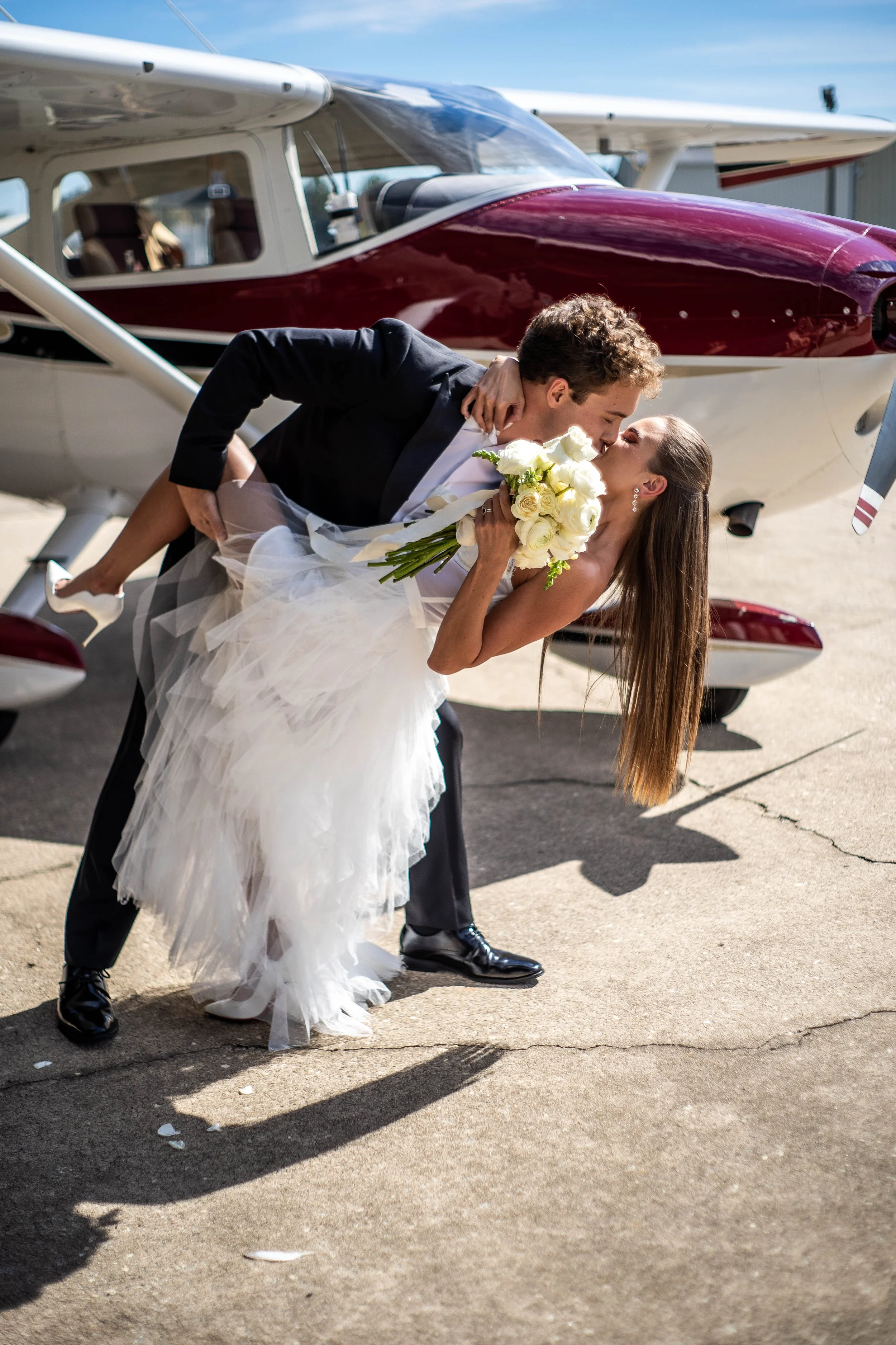 A newlywed couple sharing a kiss outdoors, with the groom dipping the bride while she holds a bouquet of white roses, in front of a small airplane.