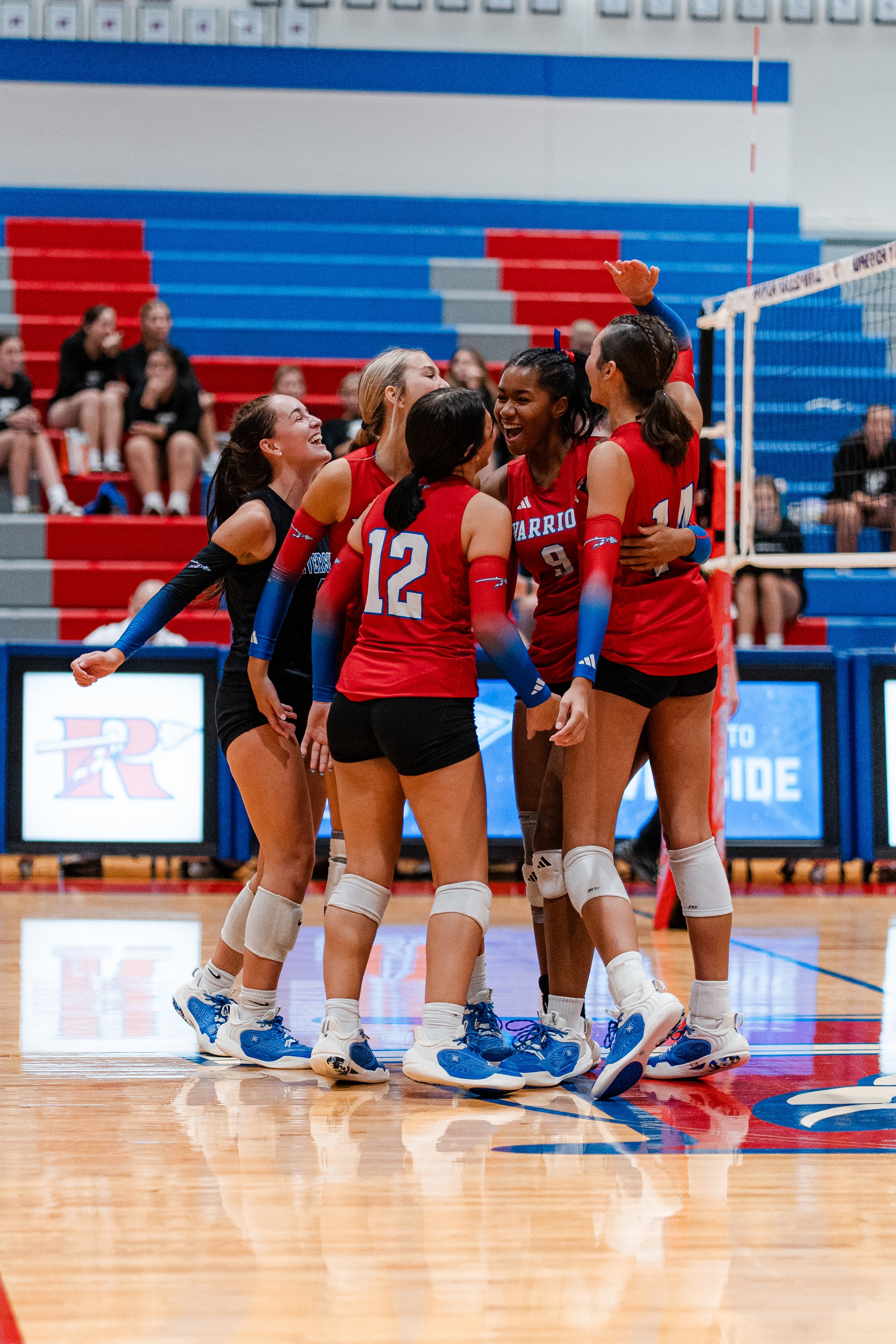 A group of female volleyball players in red and black uniforms celebrating on a gymnasium court, with spectators in the background.