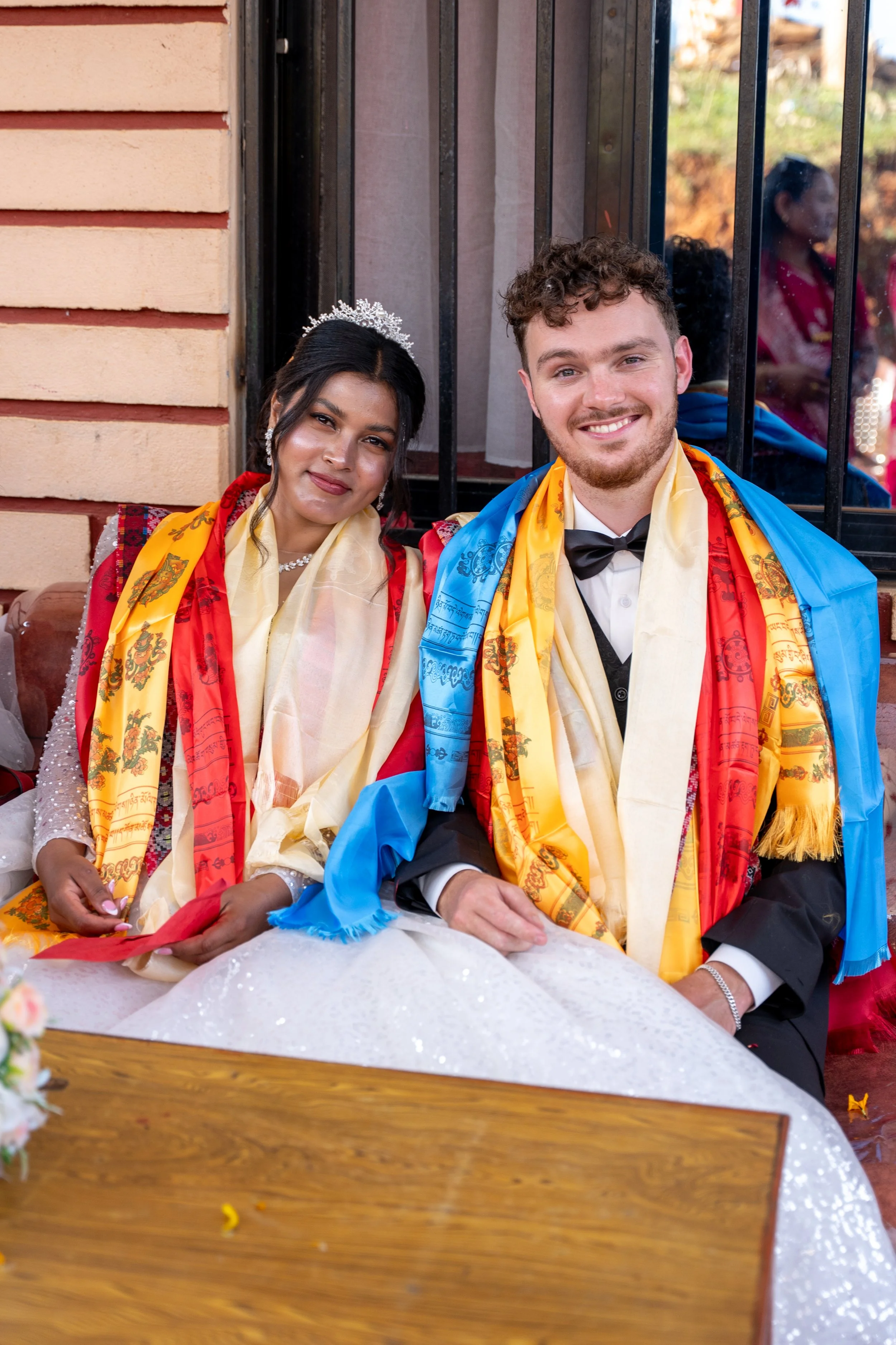 A wedding couple sitting together, dressed in formal attire, with colorful scarves draped over their shoulders, smiling at the camera.