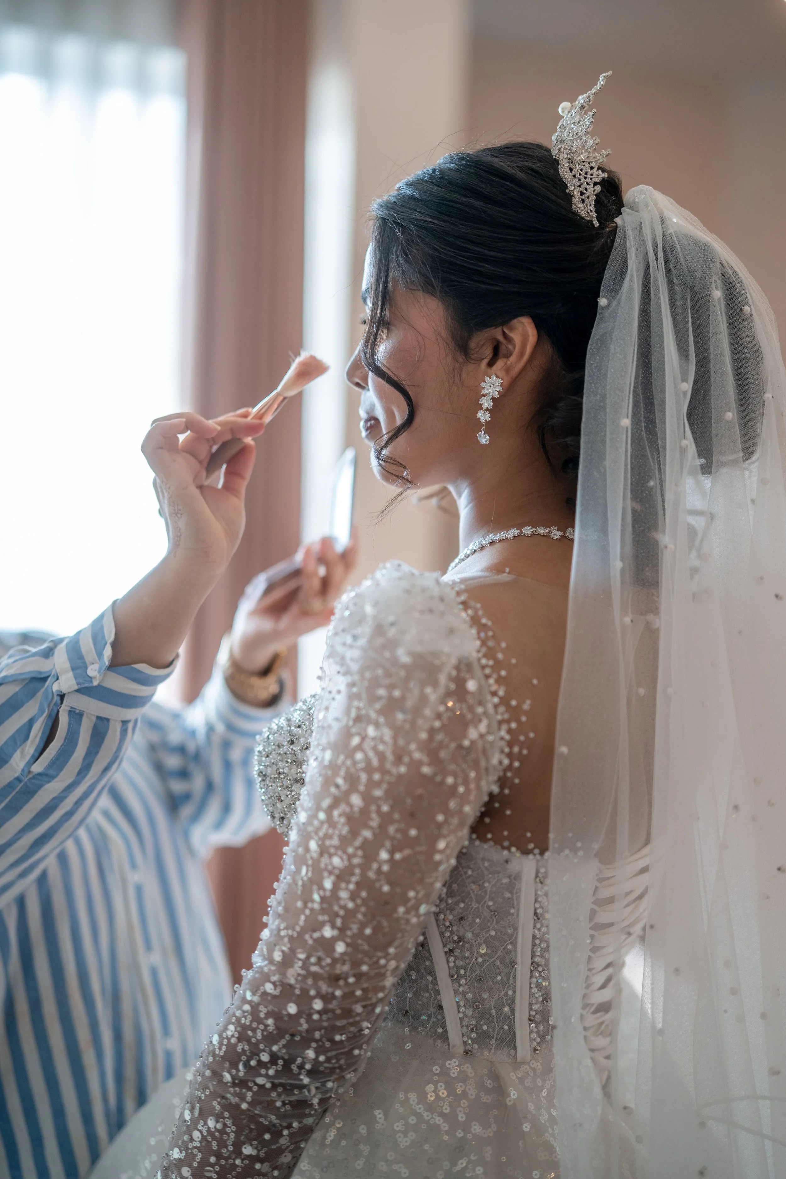 A bride with dark hair styled in waves, wearing a white wedding dress with intricate beading and lace, is having makeup applied by a makeup artist. The bride is adorned with a sparkling tiara, dangling earrings, and a matching necklace. She has a she