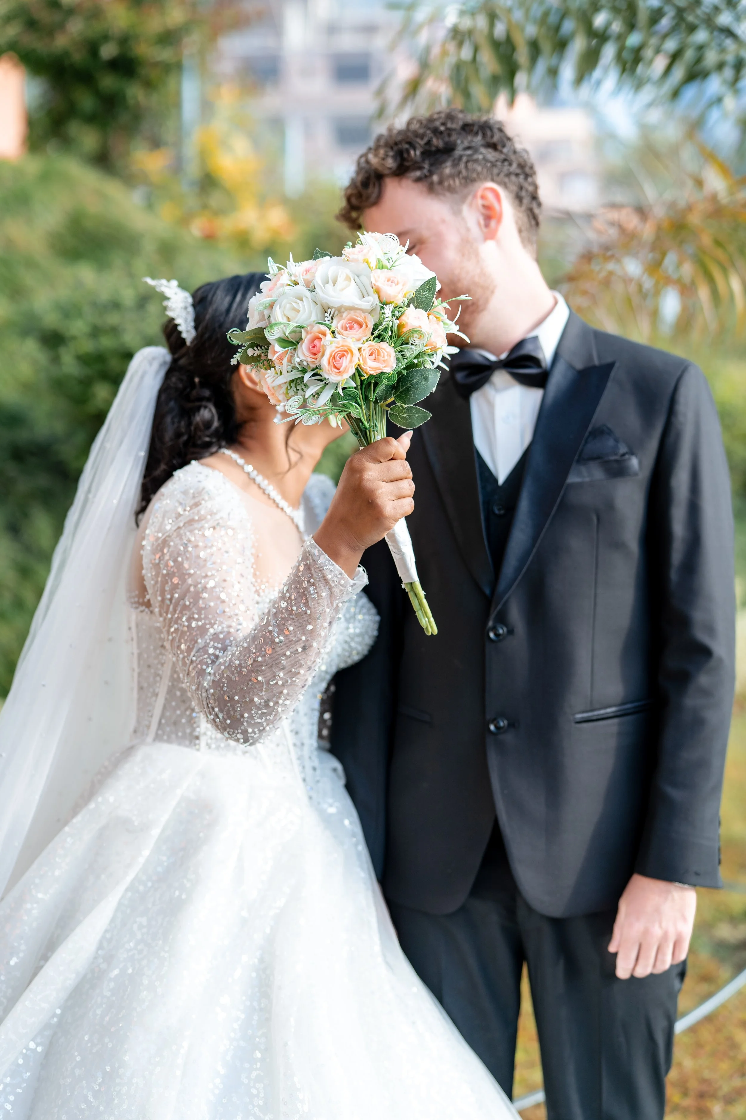 A bride and groom are exchanging a kiss outdoors, with the bride holding a bouquet of pink and white roses, wearing a sparkly white wedding gown, and the groom in a black tuxedo.