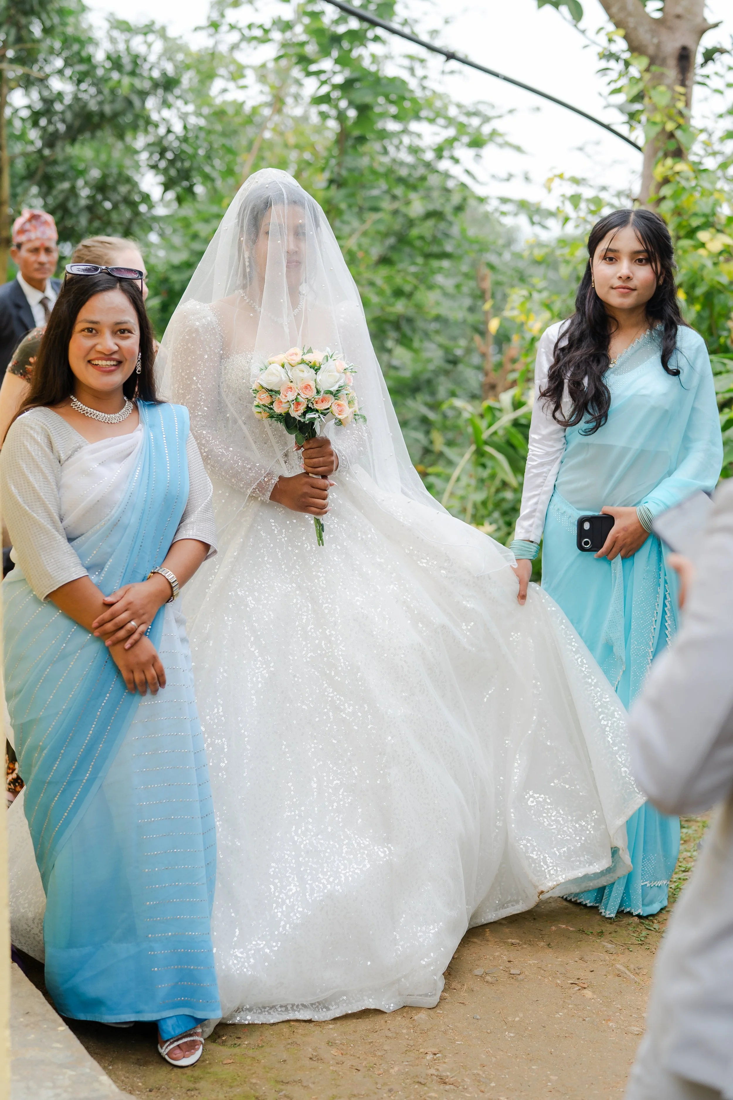 Bride in a wedding gown holding a bouquet, flanked by two women in blue traditional dresses, outdoors with green trees in the background.