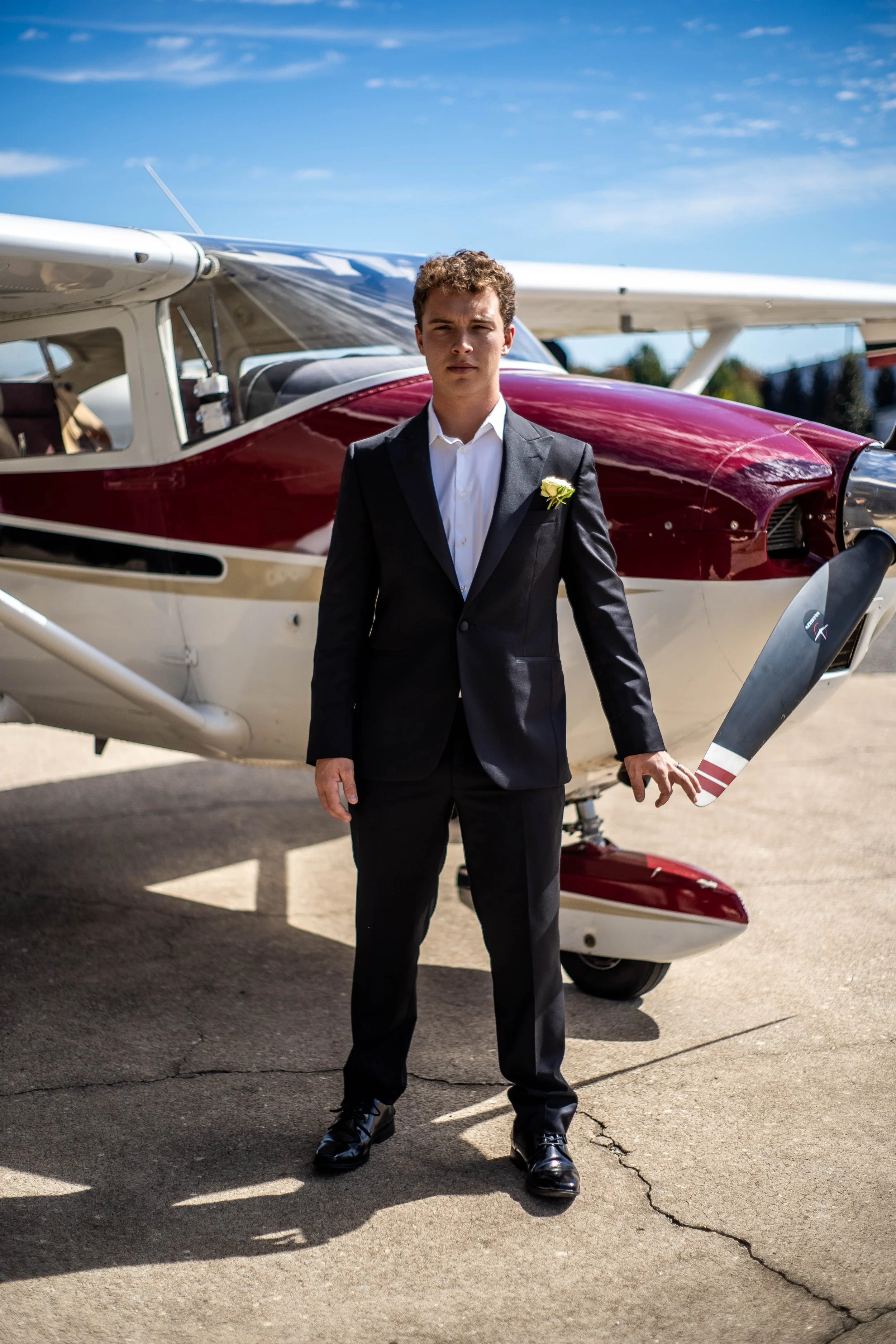 Young man in a black suit standing next to a small airplane, outdoors on a sunny day.
