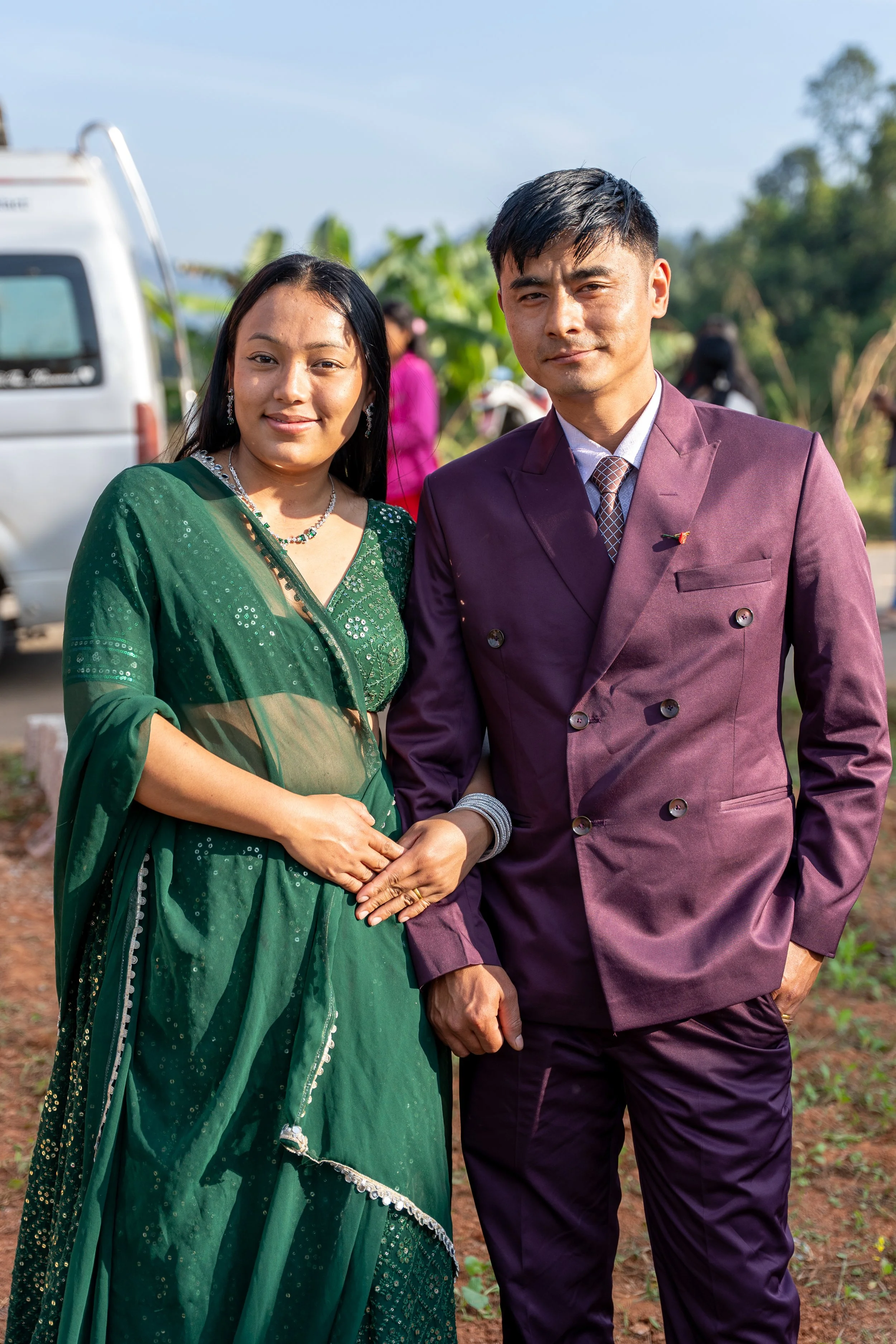 A man and woman posing outdoors, holding hands, dressed in formal and traditional attire.