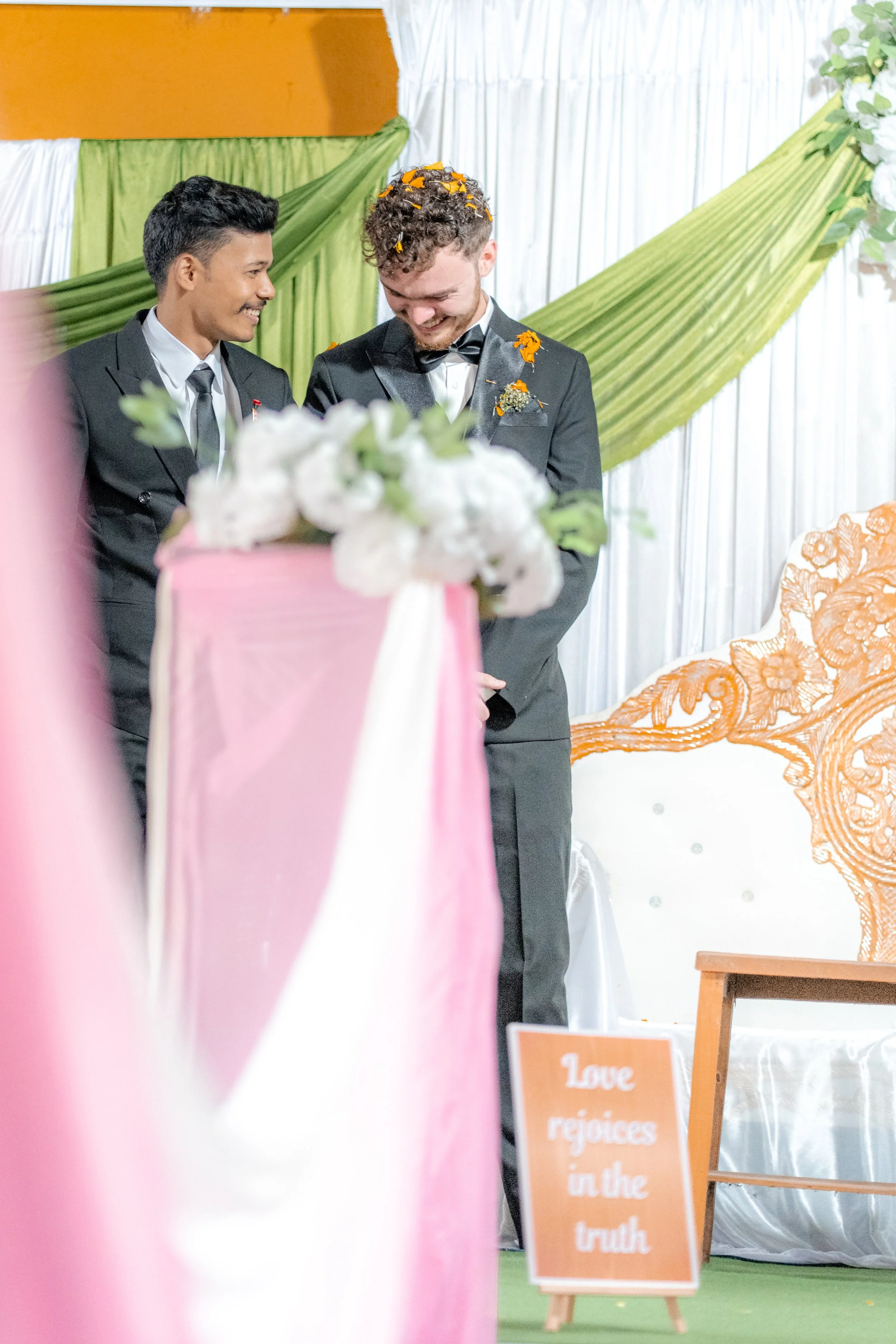 Two men in suits smiling and looking at each other at a wedding ceremony, with green and white drapery and a wooden sign that says 'Love rejoices in the truth' in the background.