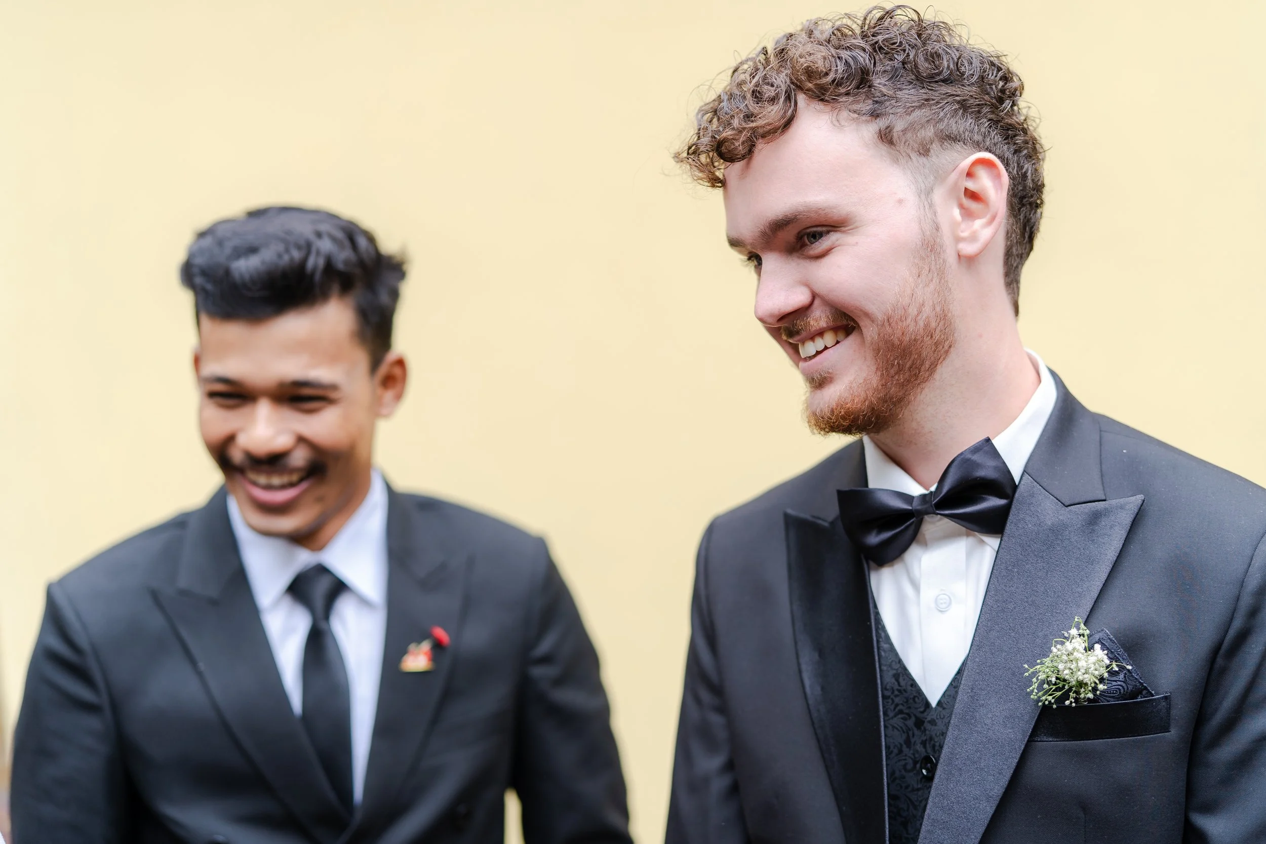Two men in black tuxedos with bow ties, smiling and looking at each other, standing against a light yellow background.