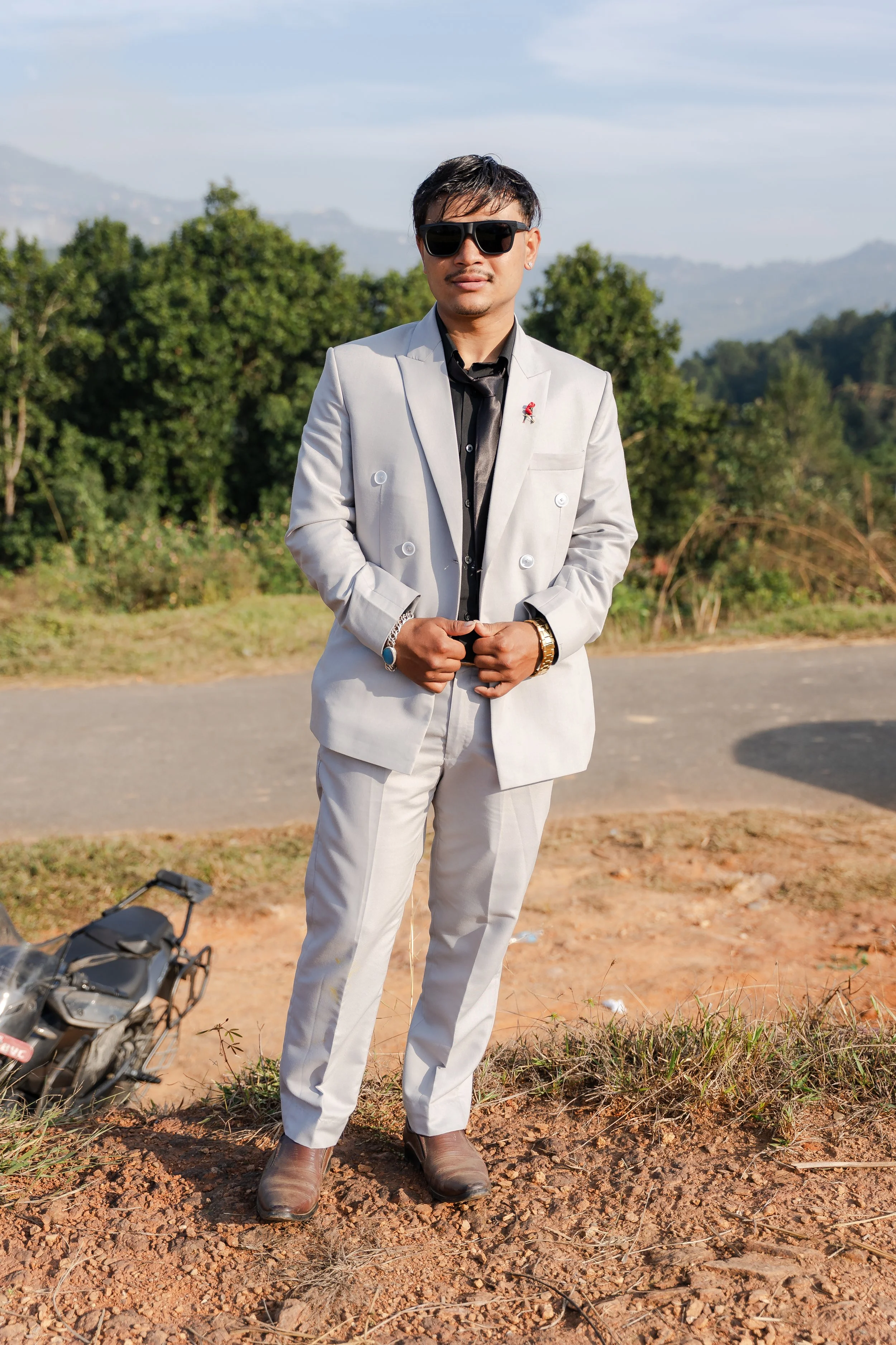 A man standing outdoors on a dirt patch, wearing a light gray suit, black shirt, and sunglasses, with green trees and mountains in the background.