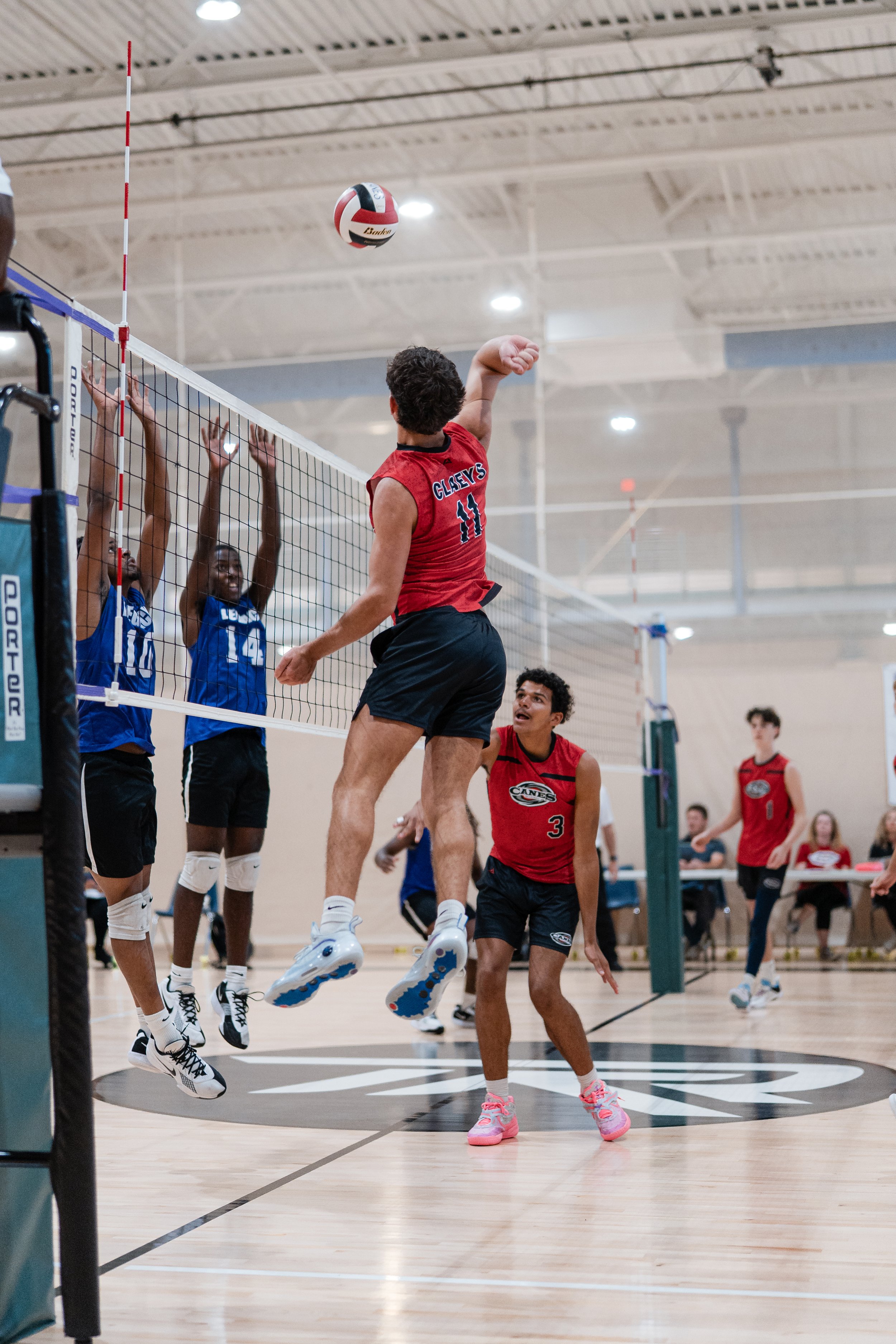 Indoor volleyball game with players jumping near the net, two players from the team in blue attempting to block, and a player in red spiking the ball.