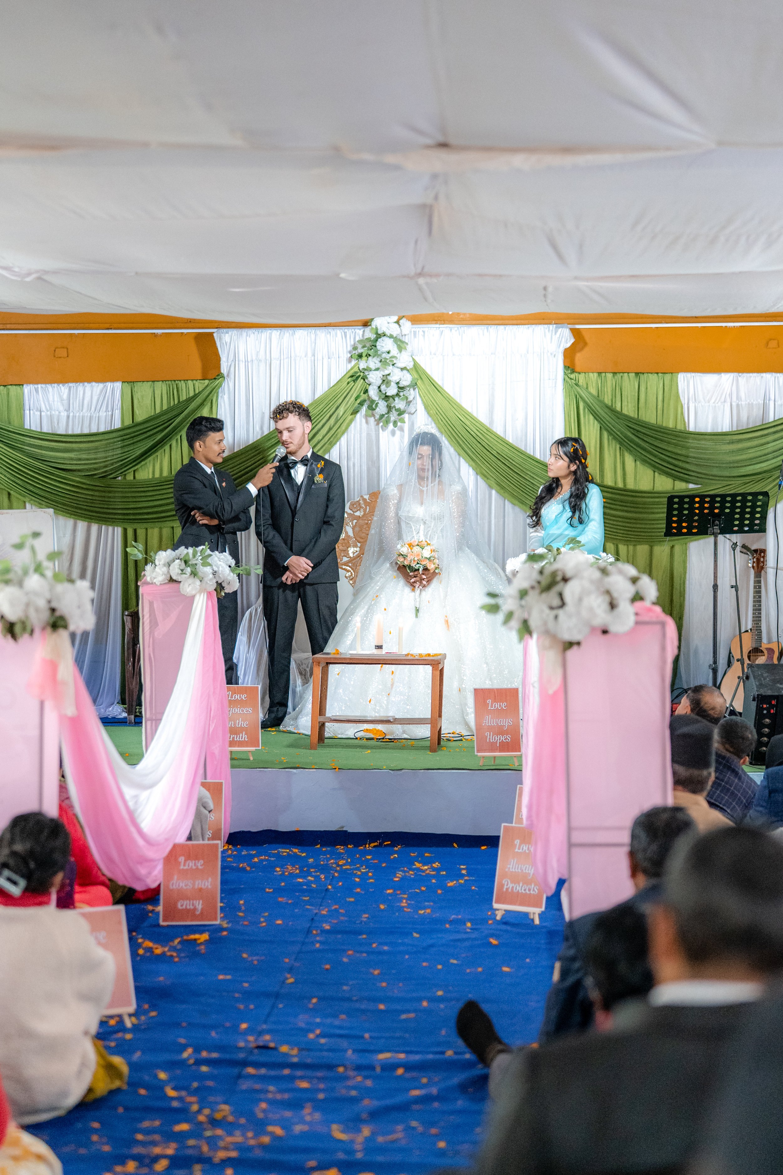 A wedding ceremony with a bride in a white gown and veil, and a groom in a black tuxedo, on a decorated stage with green and white drapes, and floral arrangements. Two individuals are standing on either side, one speaking into a microphone.