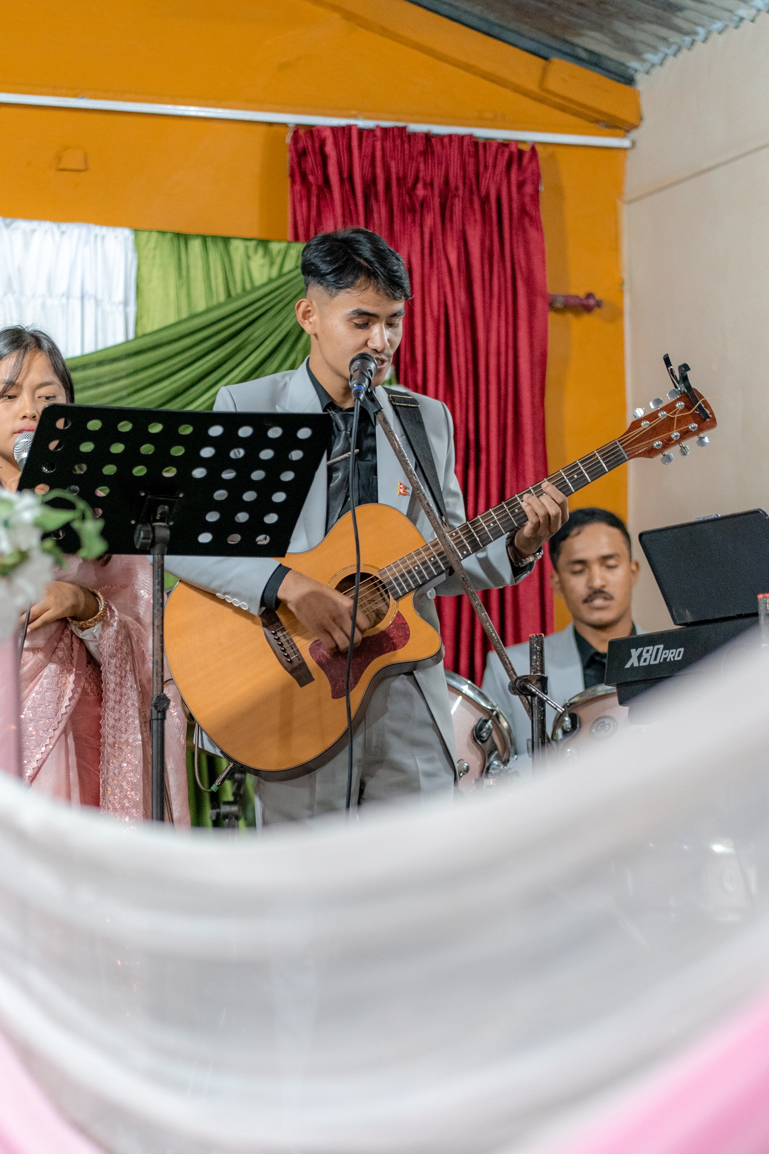 Musicians performing at a concert, with a male guitarist and singer in a grey suit prominently in front, playing an acoustic guitar and singing into a microphone.