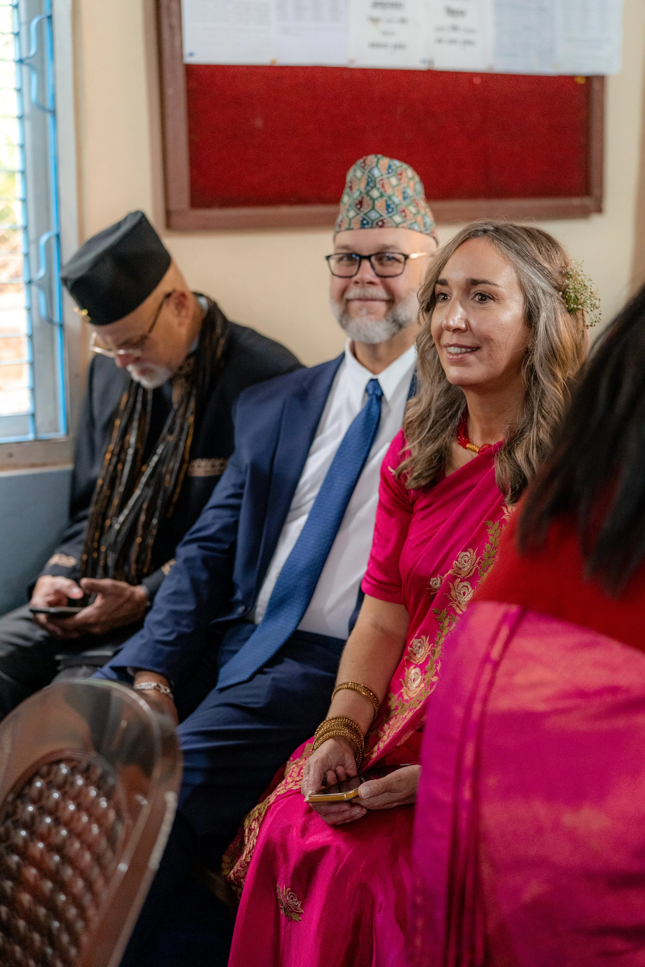 People in traditional South Asian attire sitting in a row, attending an event inside a room with a red bulletin board and a window with metal bars in the background.