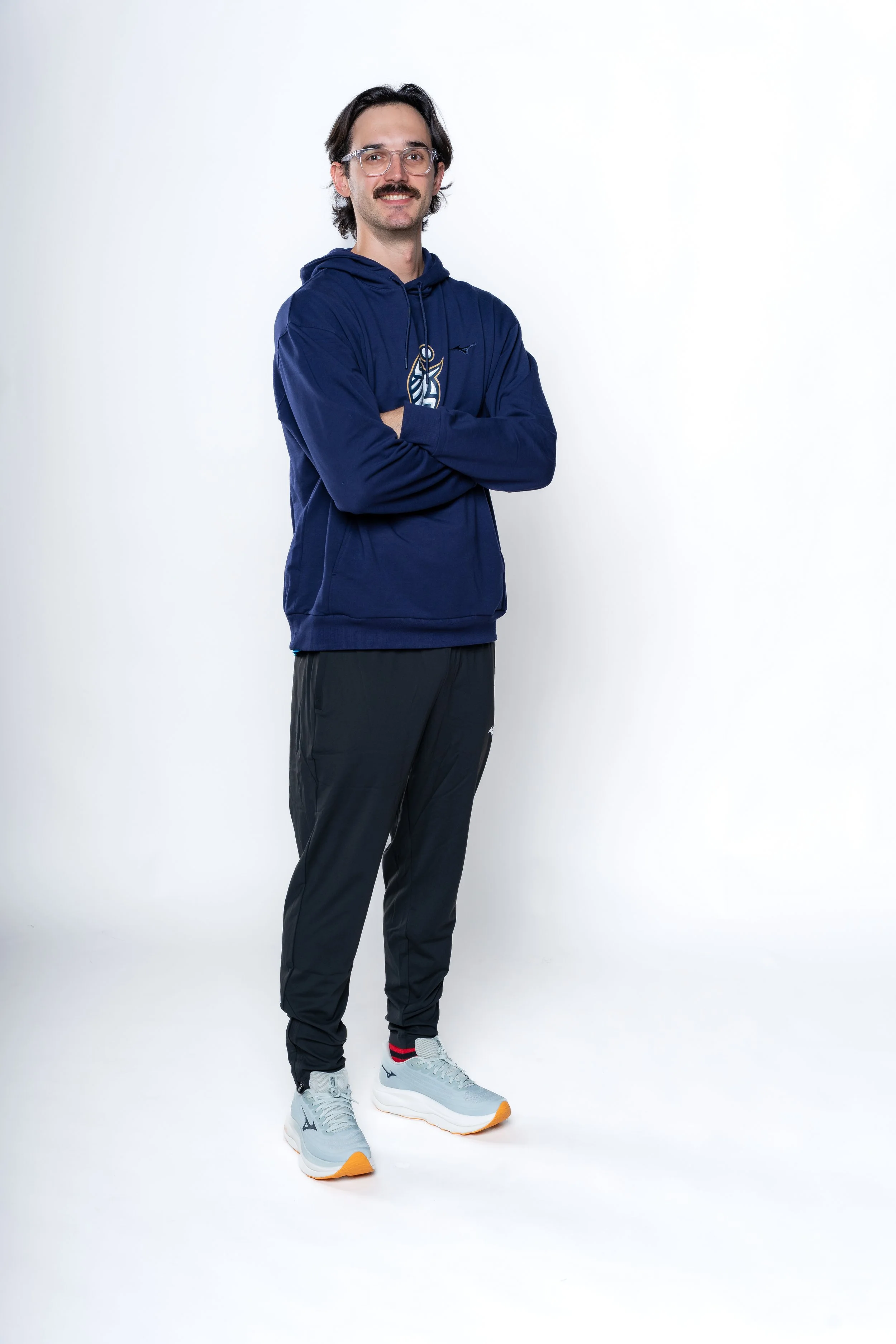 A man with glasses and a mustache wearing a navy blue hoodie, black athletic pants, and gray sneakers with orange soles, standing with arms crossed against a white background.