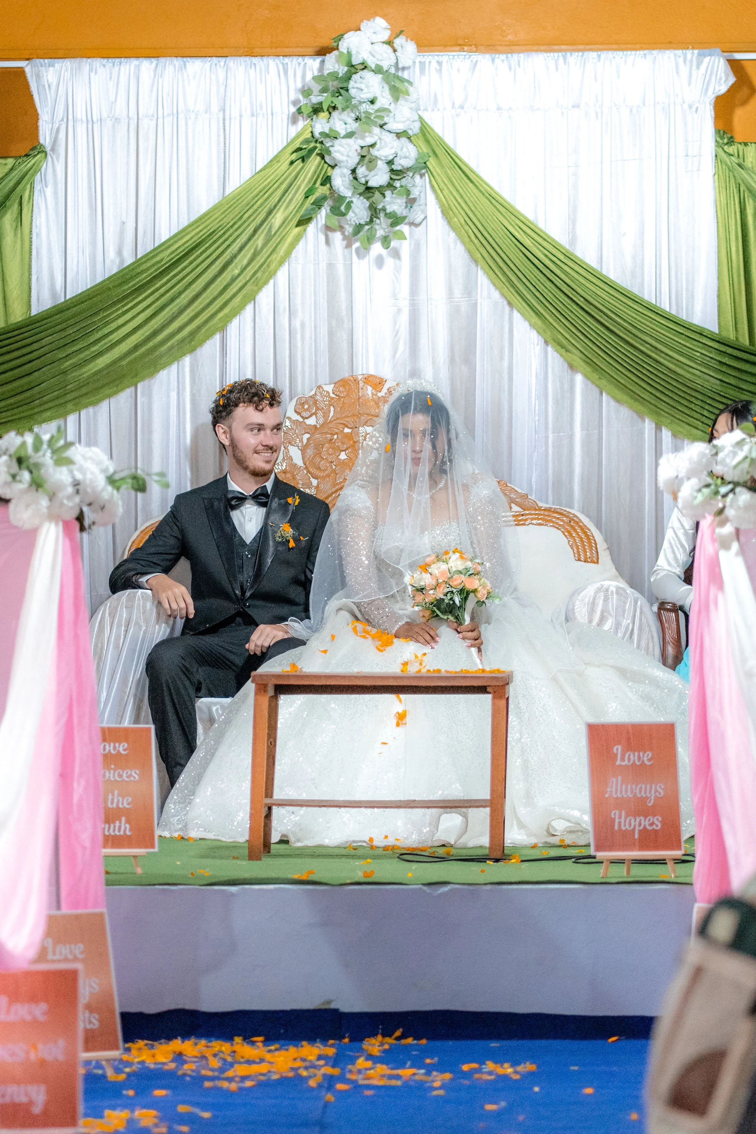 A bride and groom sitting on a decorated stage during a wedding ceremony, with flowers, draped fabric, and signs that say "Love Always Hopes."