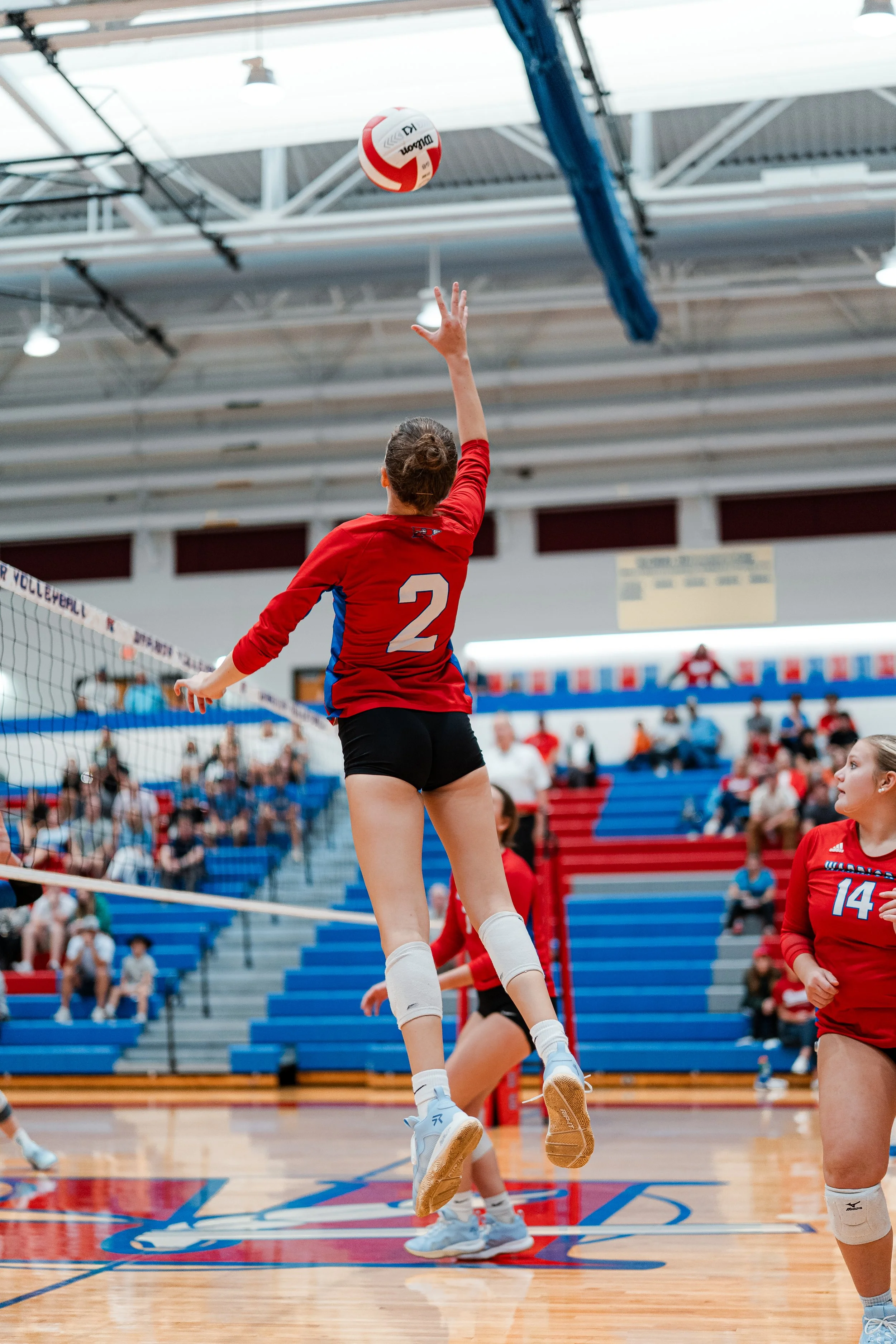 A volleyball player in a red jersey and black shorts jumping to hit the ball at an indoor volleyball court with spectators watching from the stands.