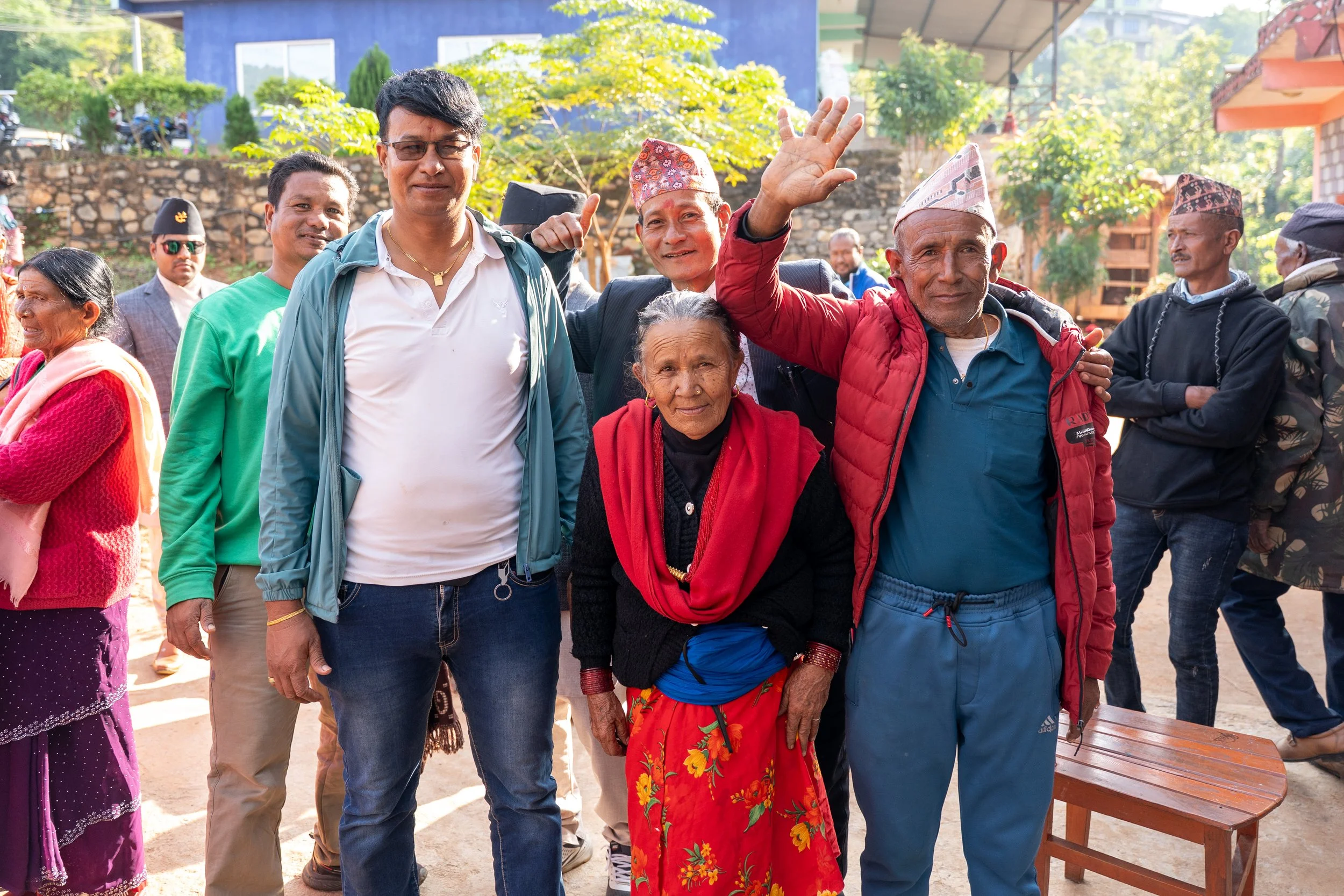 A group of Nepali people, including an elderly woman and several men, standing outdoors with trees and buildings in the background. They are smiling and waving, with sunlight illuminating the scene.