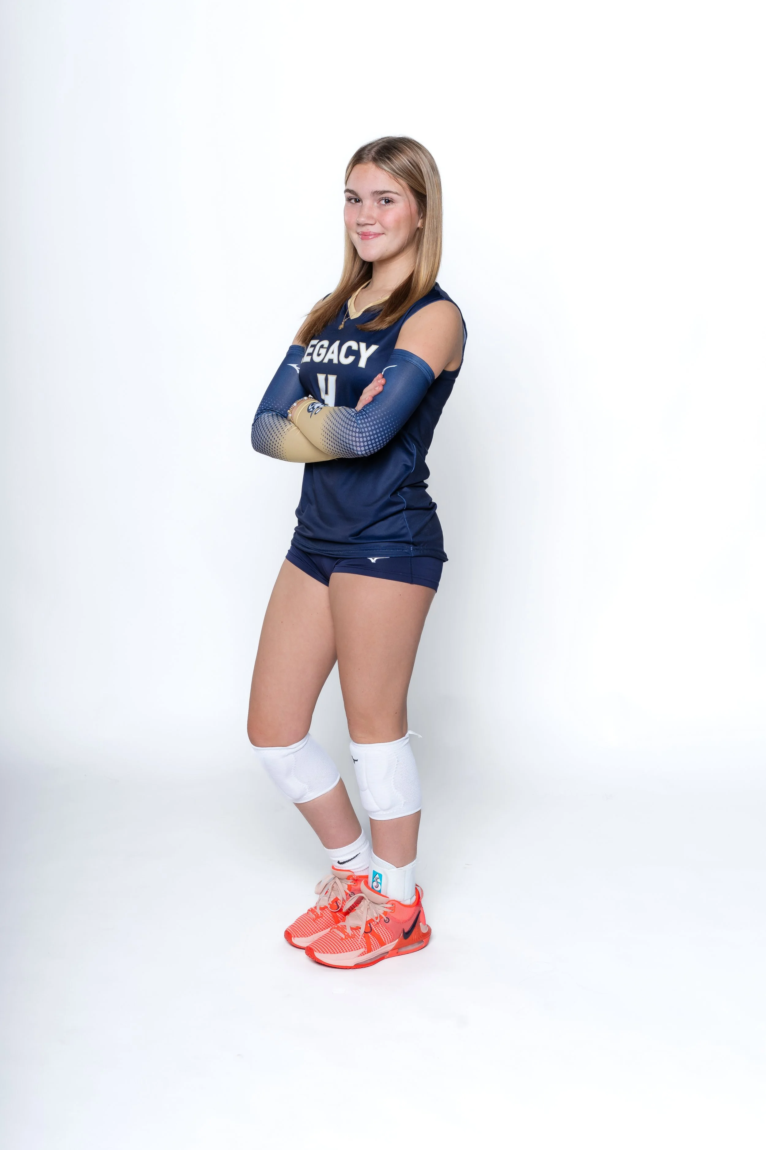 A young female volleyball player wearing a navy blue uniform with white and gold accents, knee pads, bright orange athletic shoes, and pose with her arms crossed, standing against a white background.