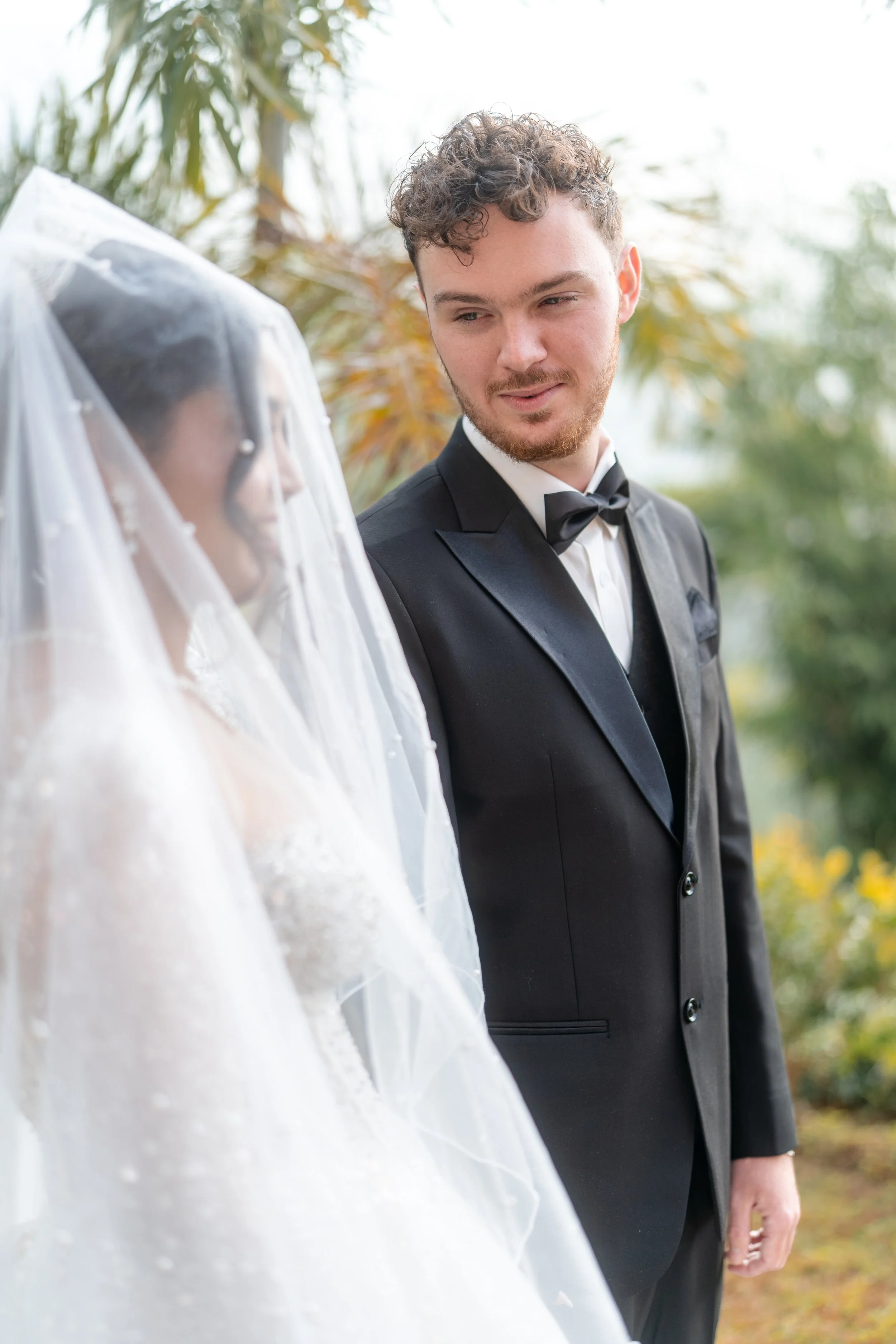 A groom in a black tuxedo and bow tie looks at a bride in a wedding dress and veil outdoors with trees and yellow flowers in the background.
