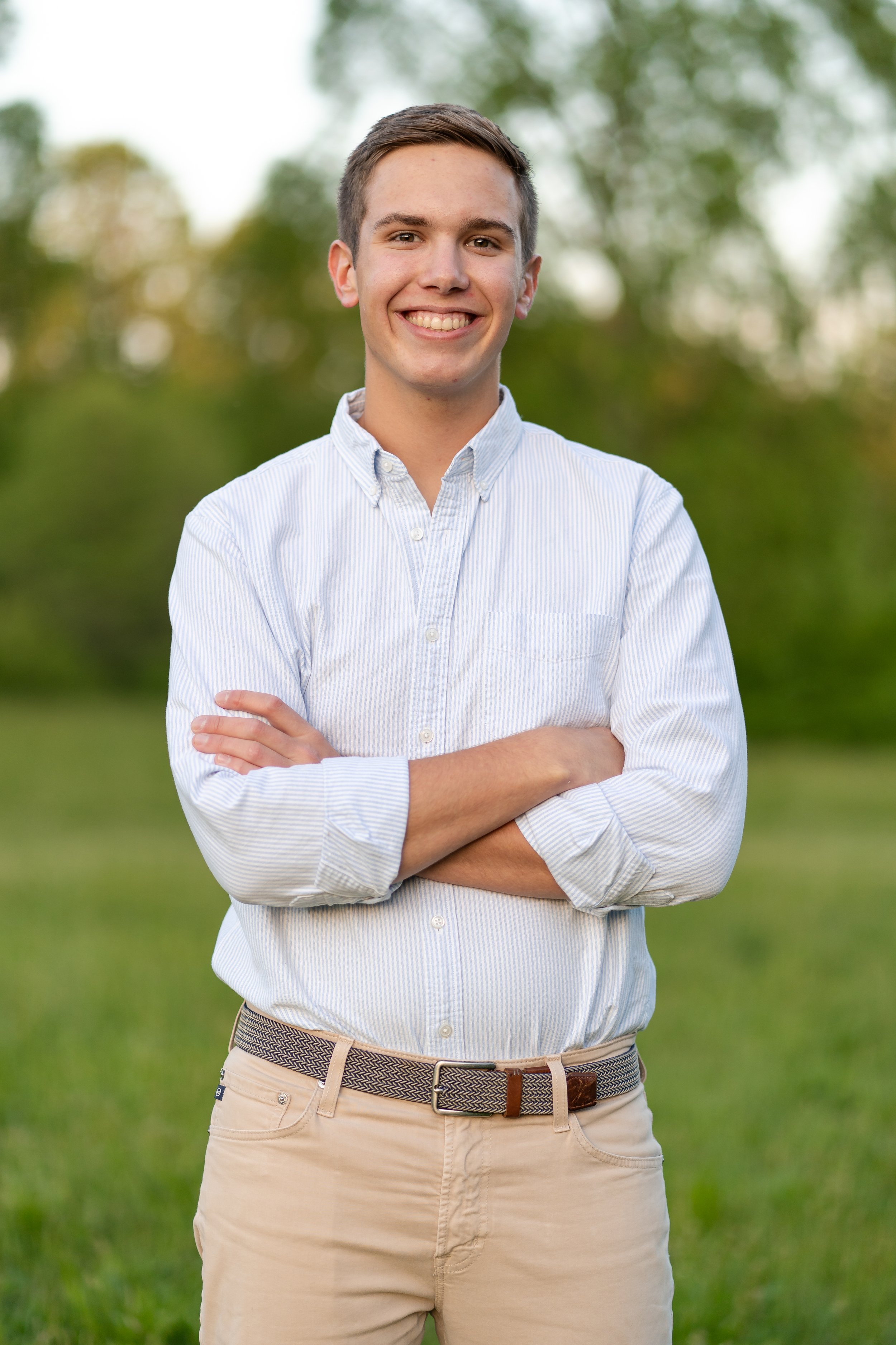 A smiling young man with short brown hair wearing a white button-up shirt with rolled-up sleeves and beige pants, standing with crossed arms outdoors on green grass with trees in the background.