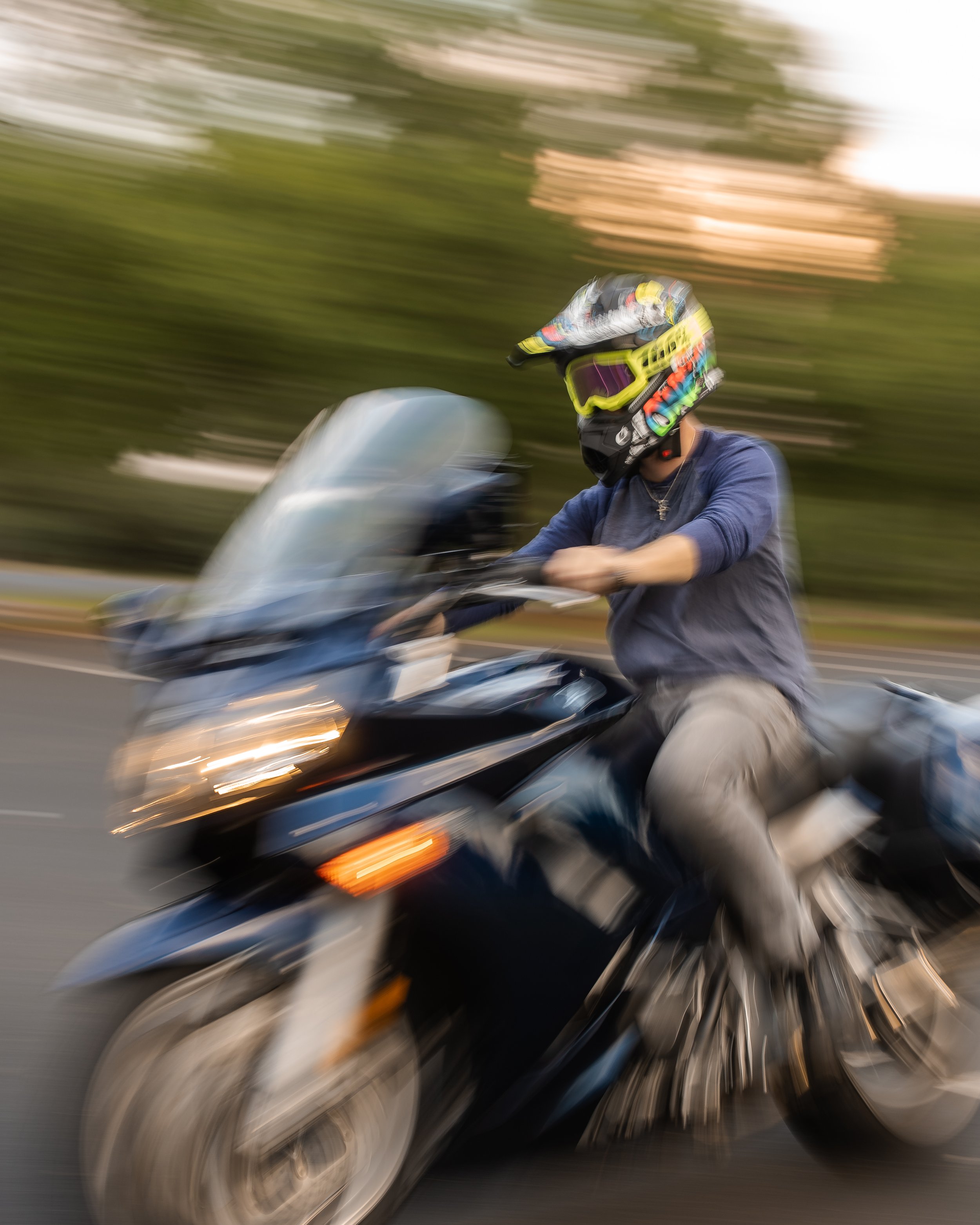 A person riding a motorcycle on a road, wearing a helmet with goggles and a mask, with a colorful helmet design. The background shows trees and it appears to be moving fast.