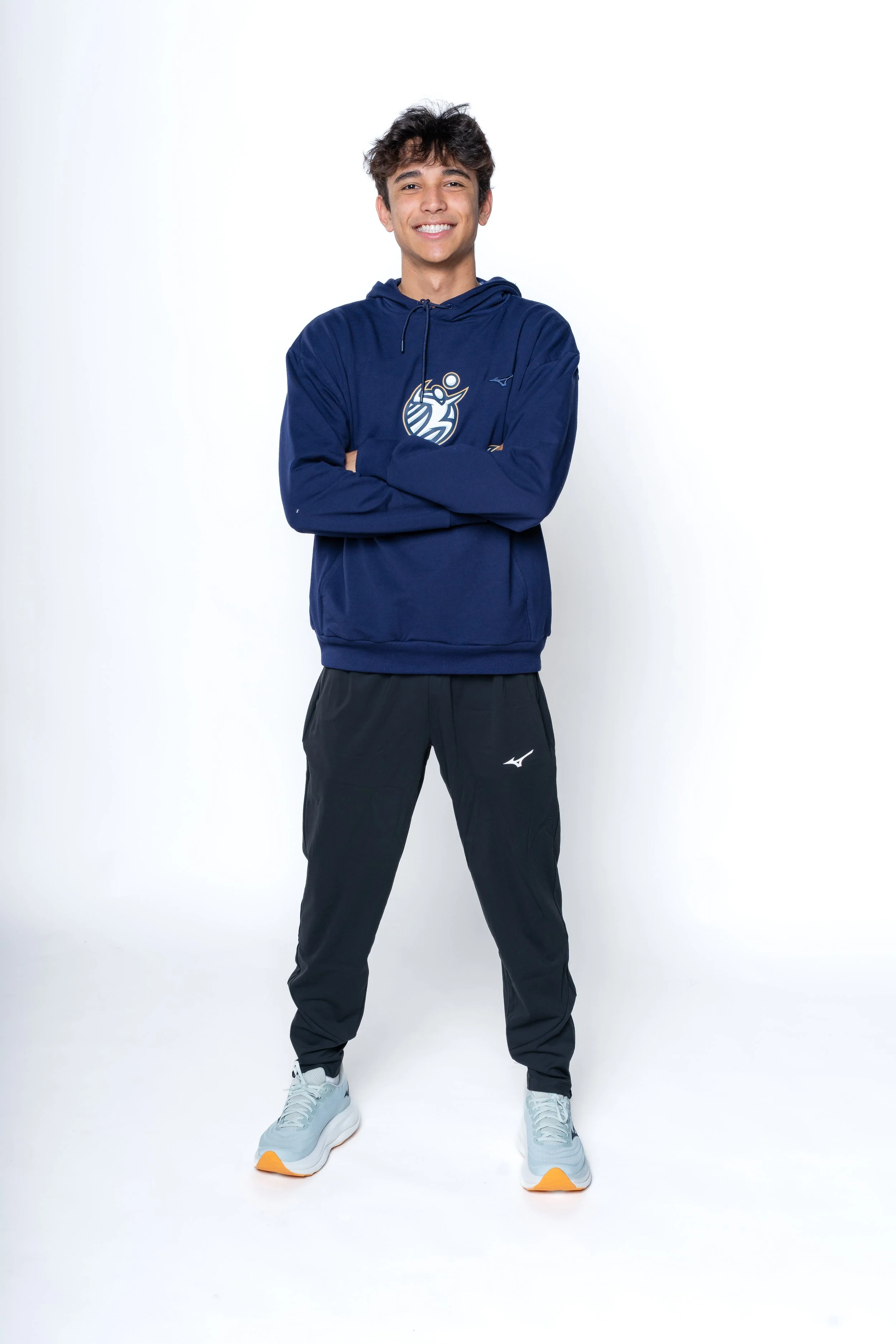 A young man standing with arms crossed, smiling, wearing a navy hoodie, black athletic pants, and gray sneakers, on a white background.