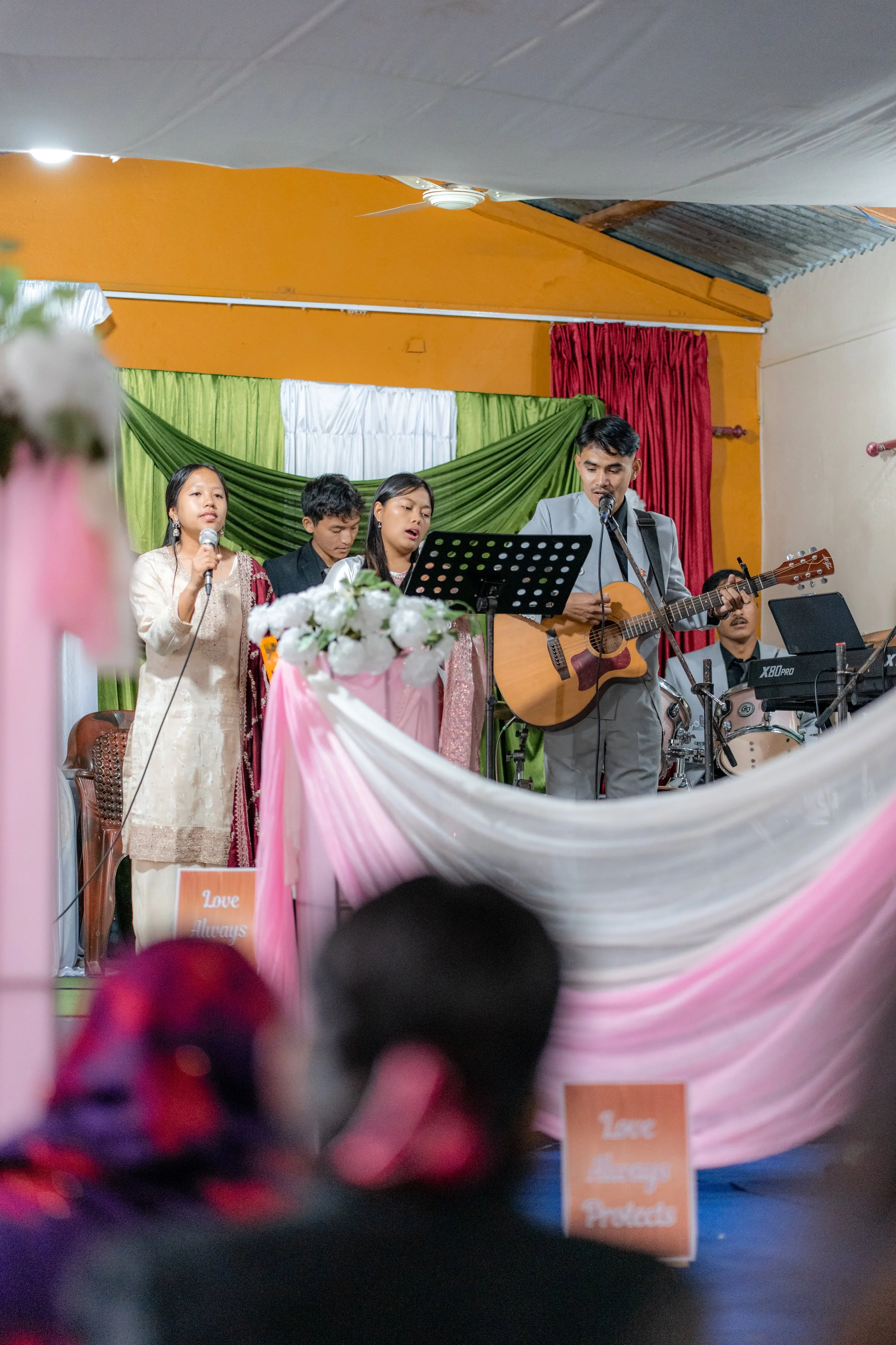 A church celebration with a live band on stage, including a singer, guitarist, and drummer, with decorative pink, white, and green drapes and signs reading "Love Always Protects."