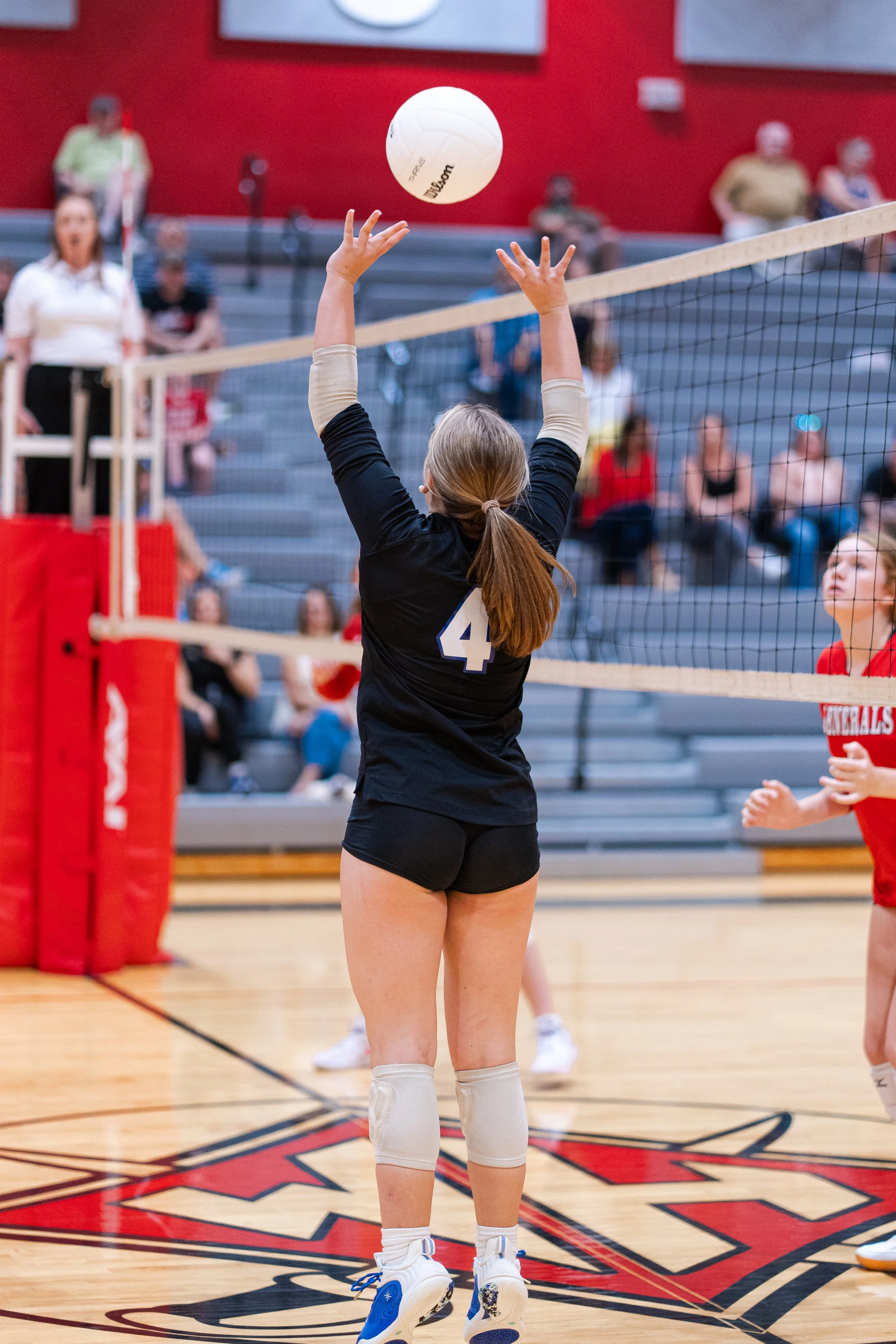 A volleyball player with jersey number 4 spiking the ball over the net during a game in a gymnasium, with spectators seated on the bleachers in the background.