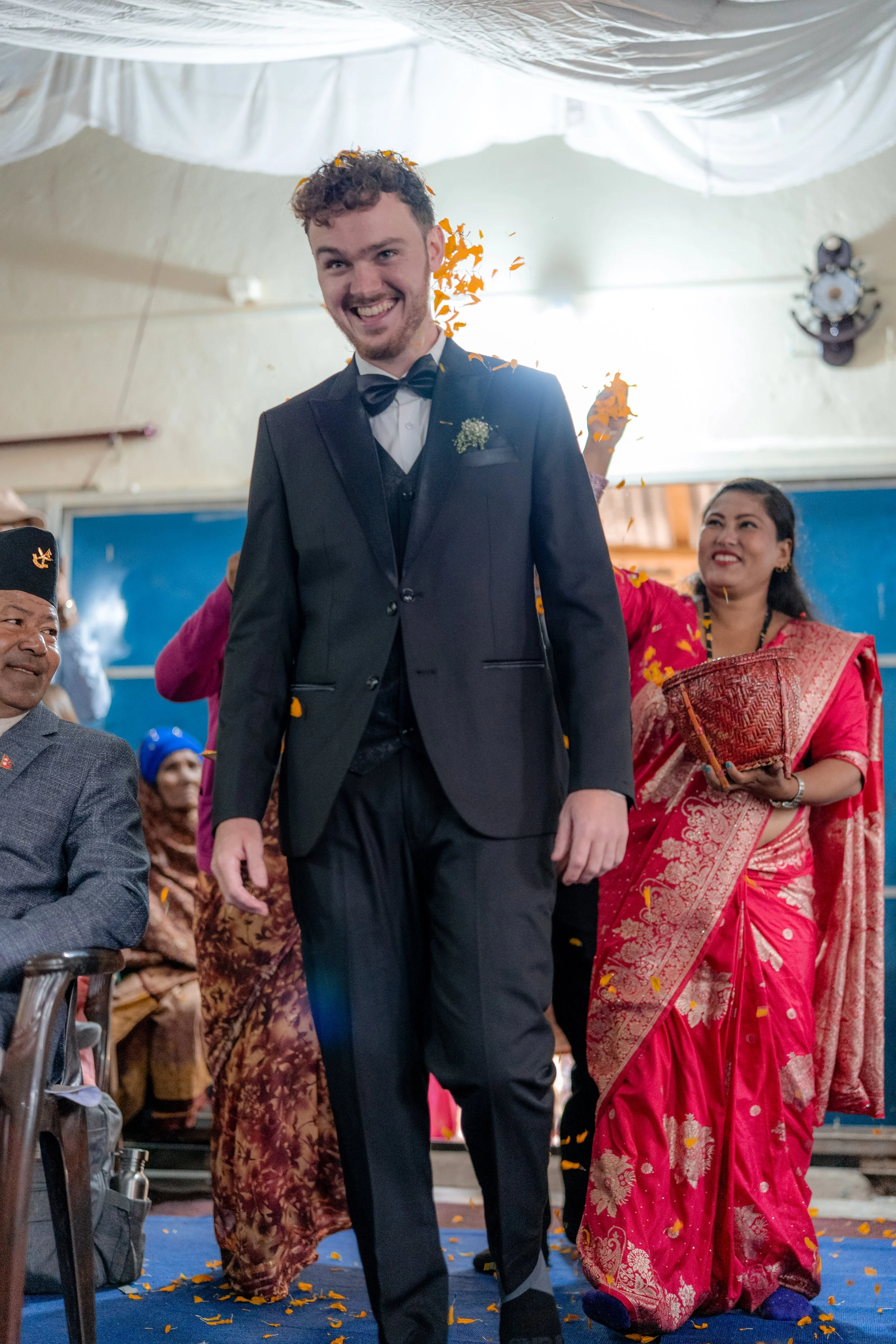 A cheerful groom in a dark suit with a bowtie walking down an aisle, surrounded by people celebrating, including a woman in a traditional red sari holding a basket, during a wedding ceremony with flower petals and confetti.