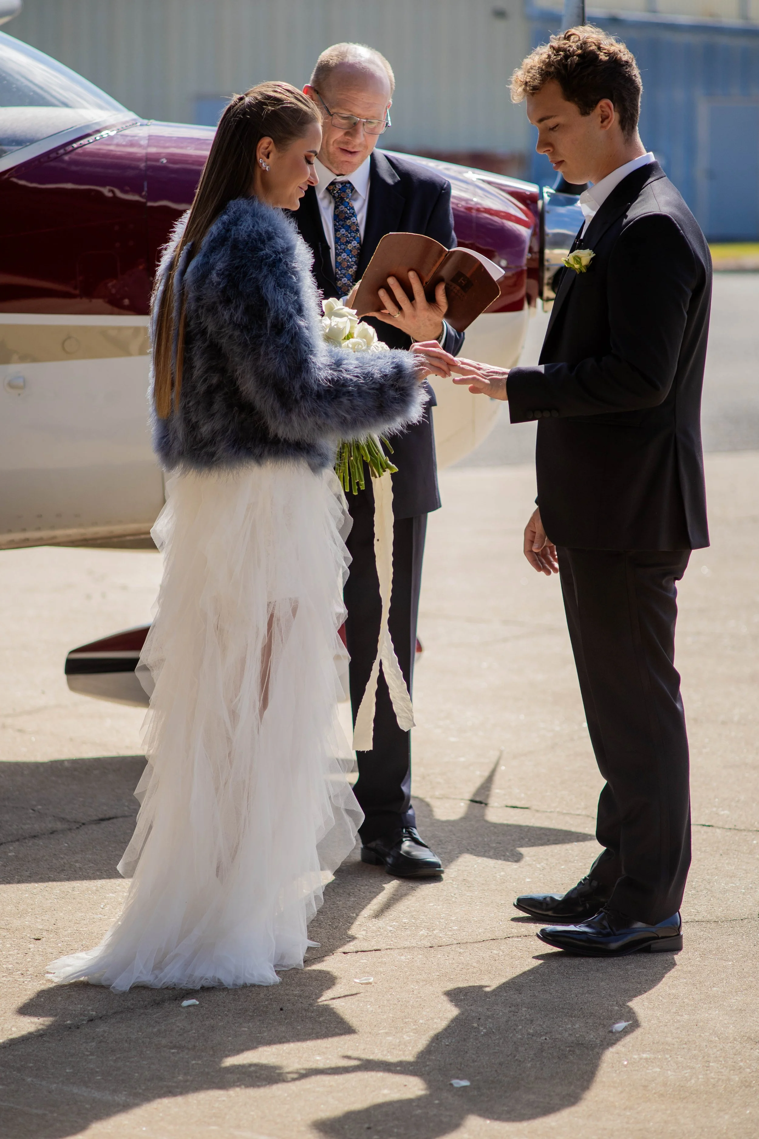A couple getting married outdoors, with an officiant reading from a book. The bride wears a white wedding dress with a faux fur jacket, and the groom is in a black tuxedo with a boutonniere. A small plane is in the background.
