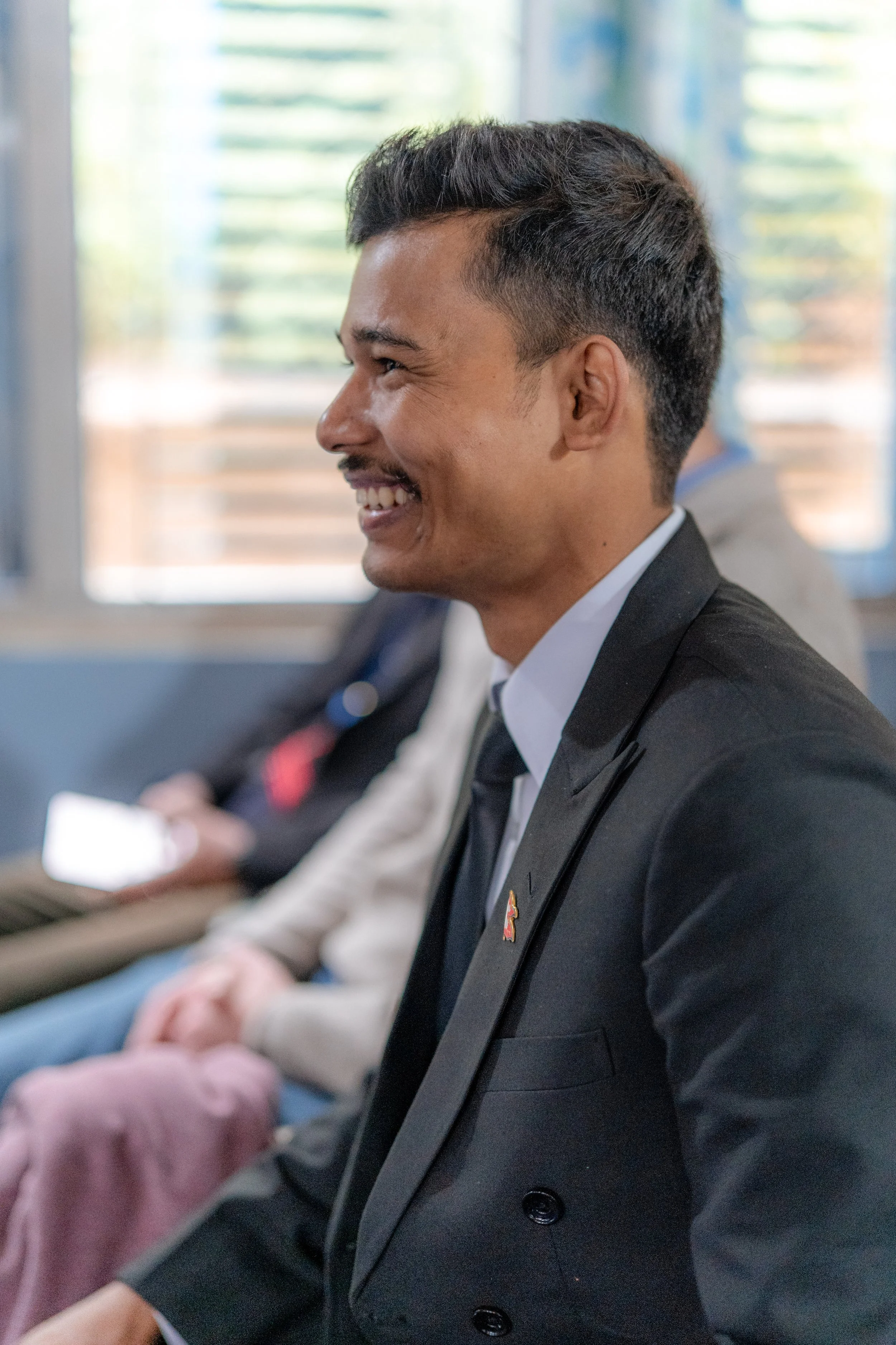 Smiling man in a black suit and tie sitting in a row with other people at a formal event.
