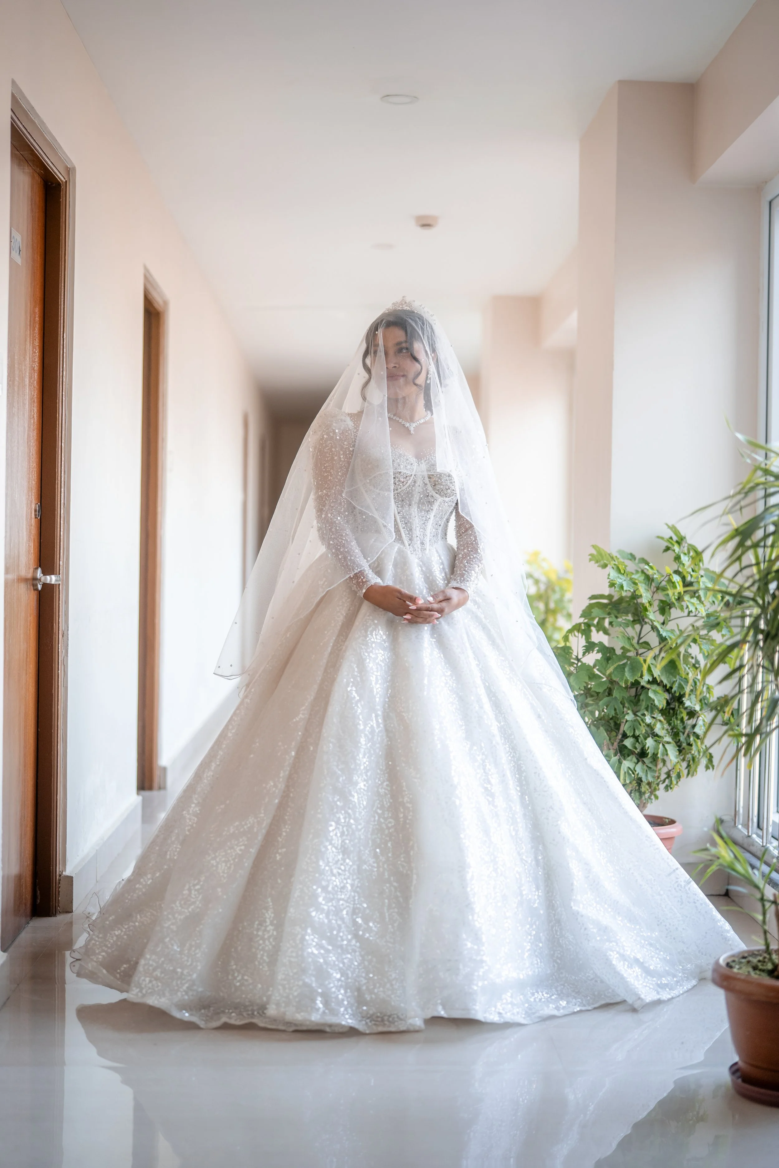 A bride in a sparkling white wedding gown with a veil standing in a well-lit hallway with potted plants.