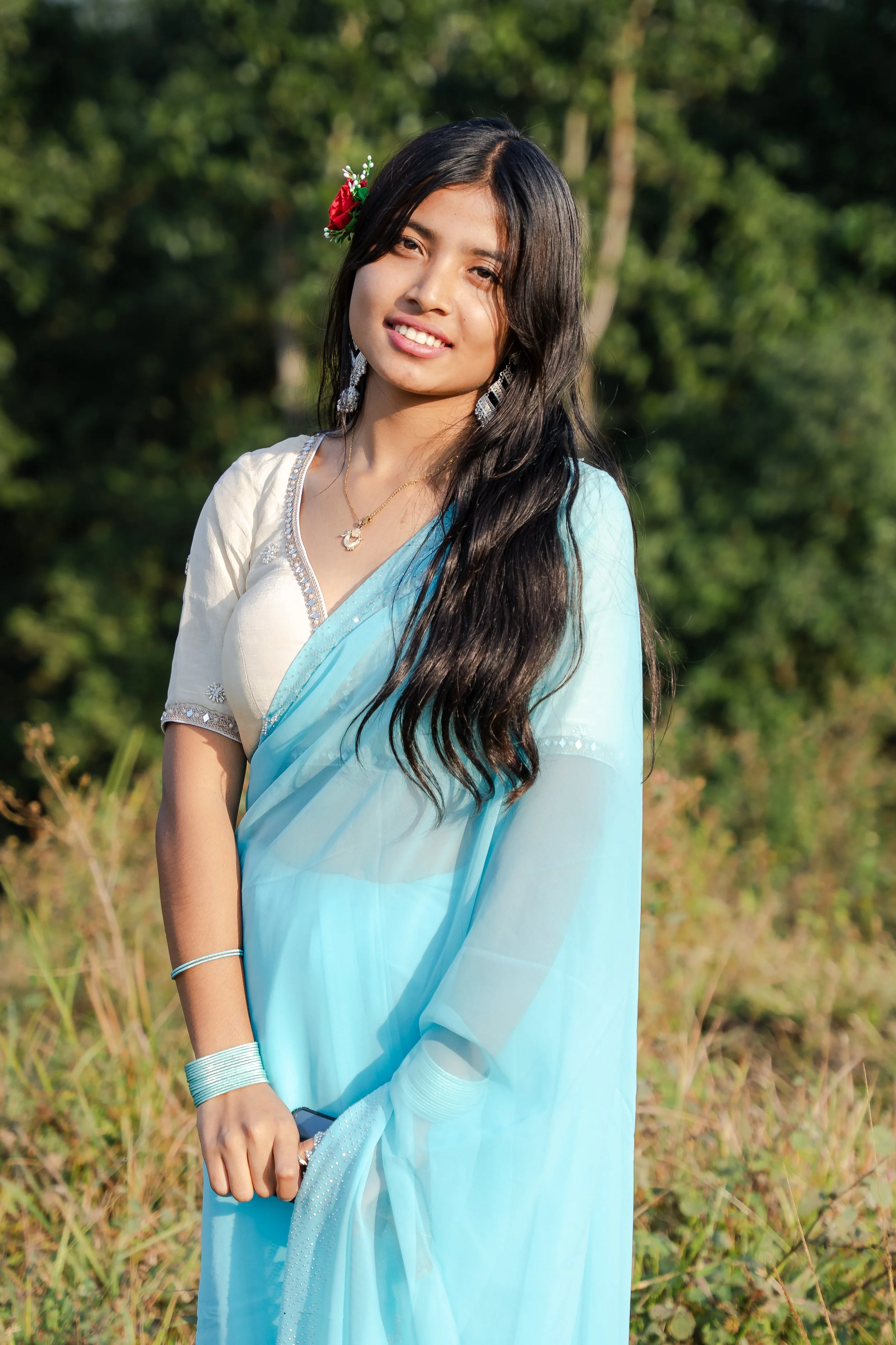 A young woman wearing a light blue saree and jewelry standing outdoors in a natural setting.