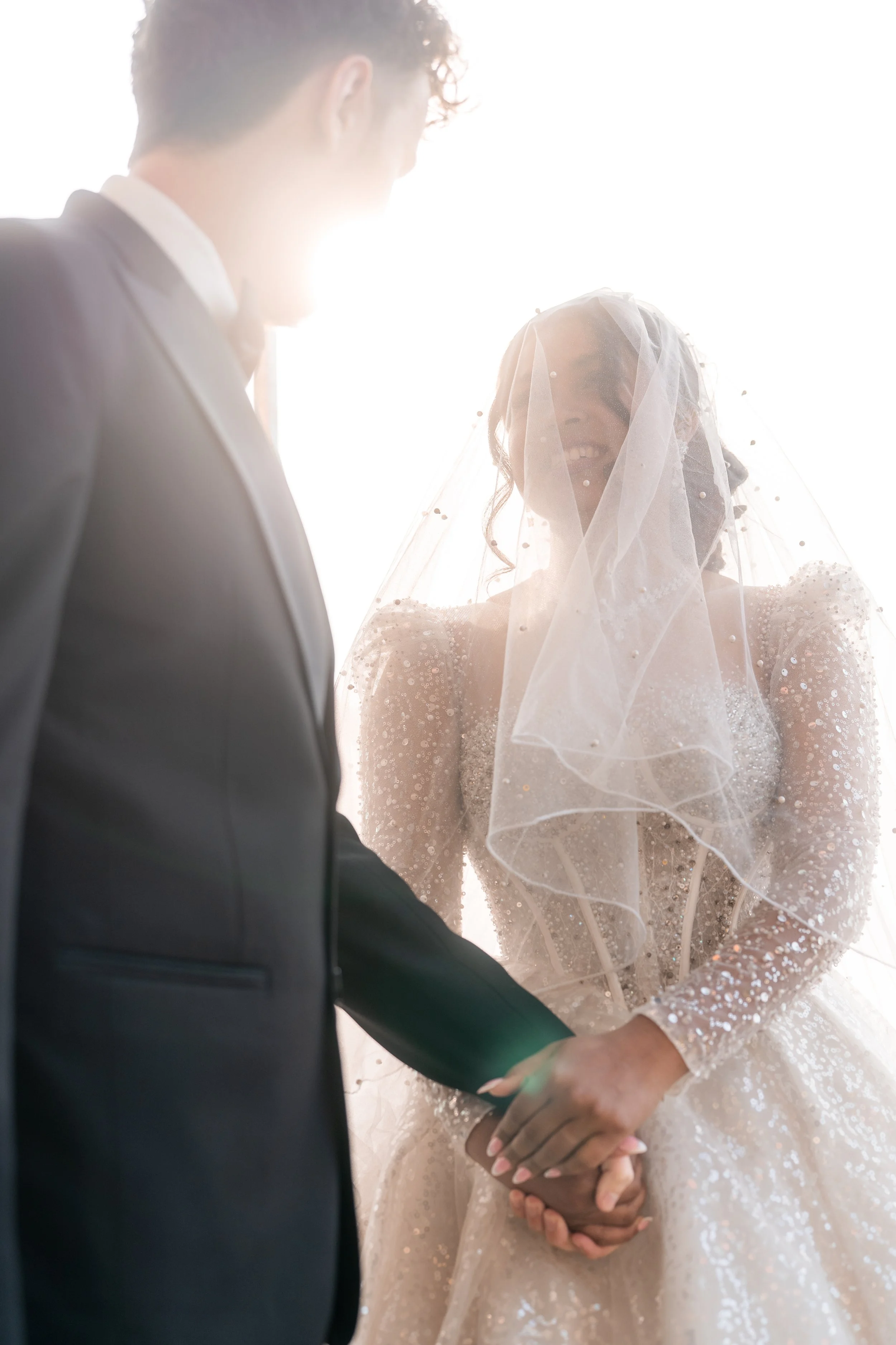 A bride and groom holding hands during their wedding ceremony, with the bride wearing a detailed, sparkly wedding dress and veil, and the groom in a dark suit. The sunlight creates a bright backlit effect.