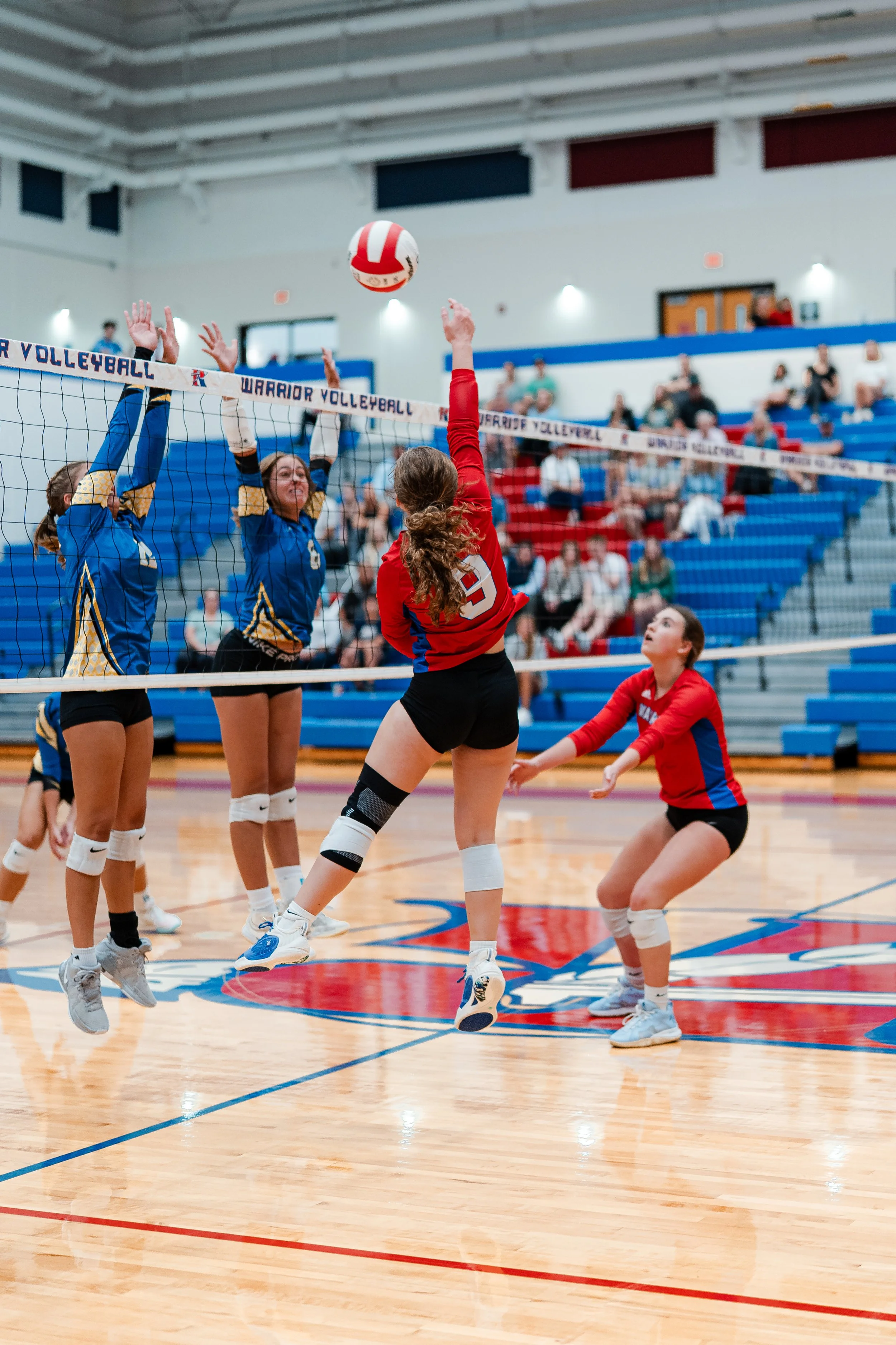 Girls playing volleyball in an indoor gym, with one girl in red jumping to spike the ball while others attempt to block.