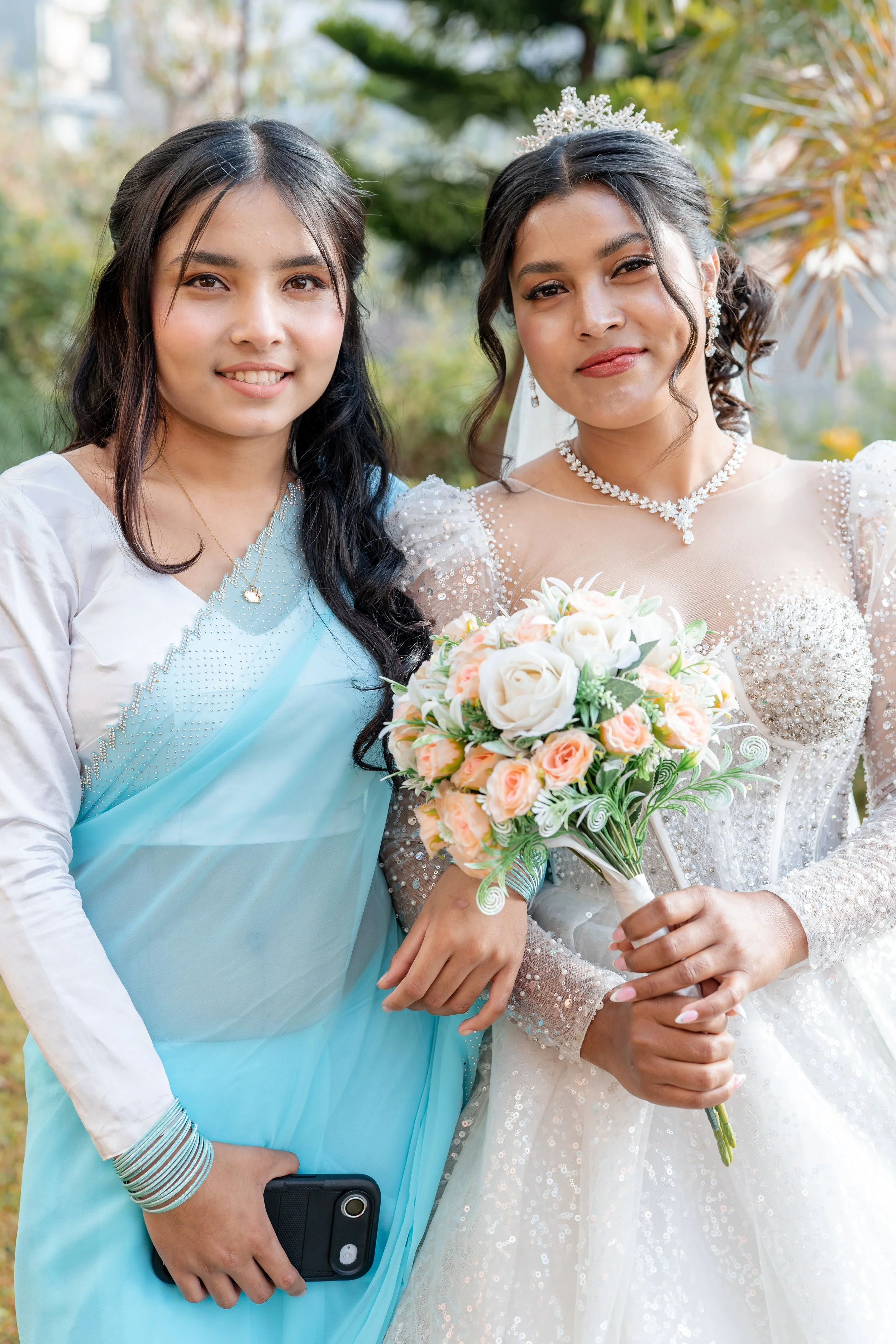 A woman in a white wedding dress holding a bouquet of flowers, standing next to a woman in a light blue sari, outdoors with greenery in the background.