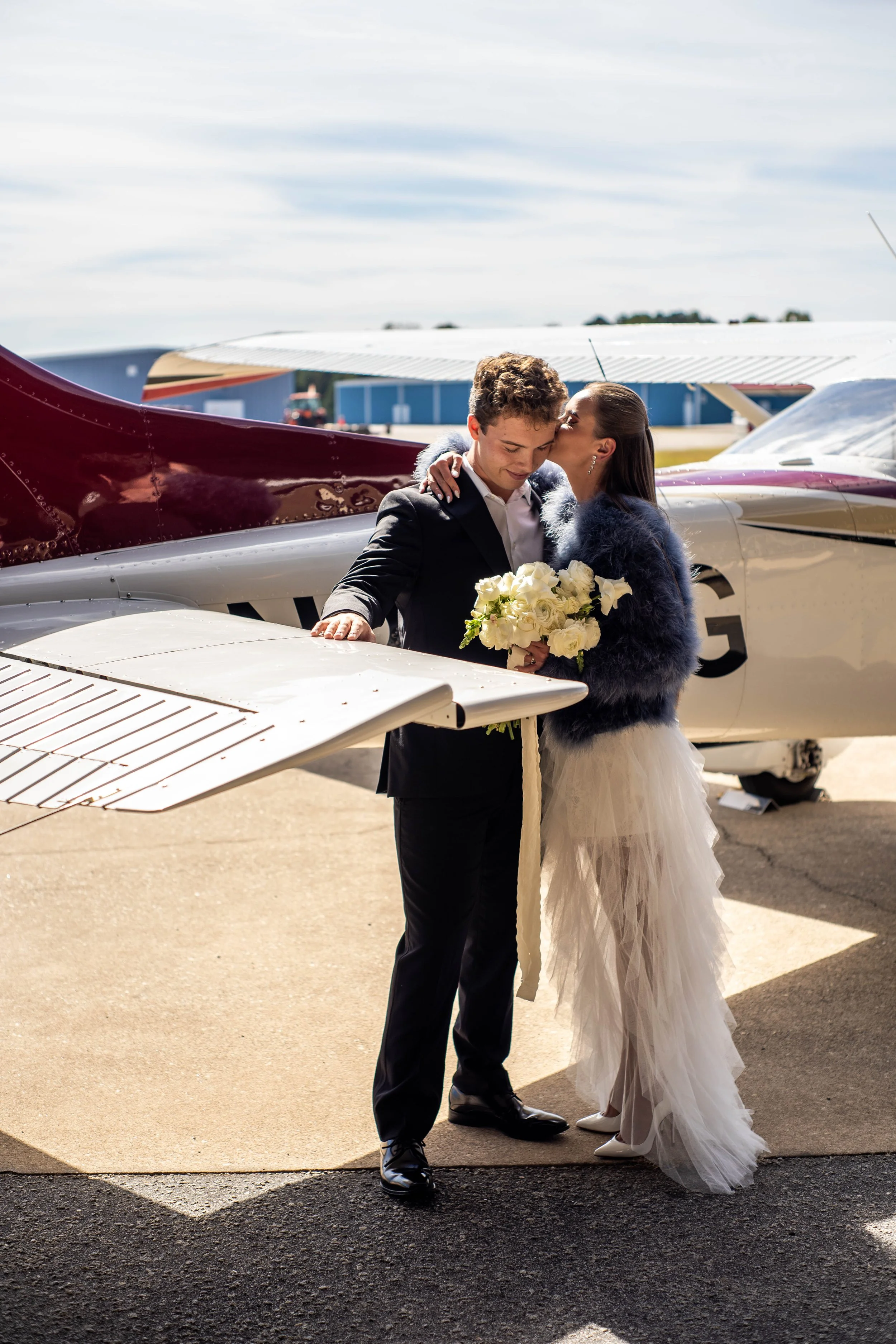 A bride and groom embrace in front of a small airplane at an airport, with the bride holding a bouquet of white roses.