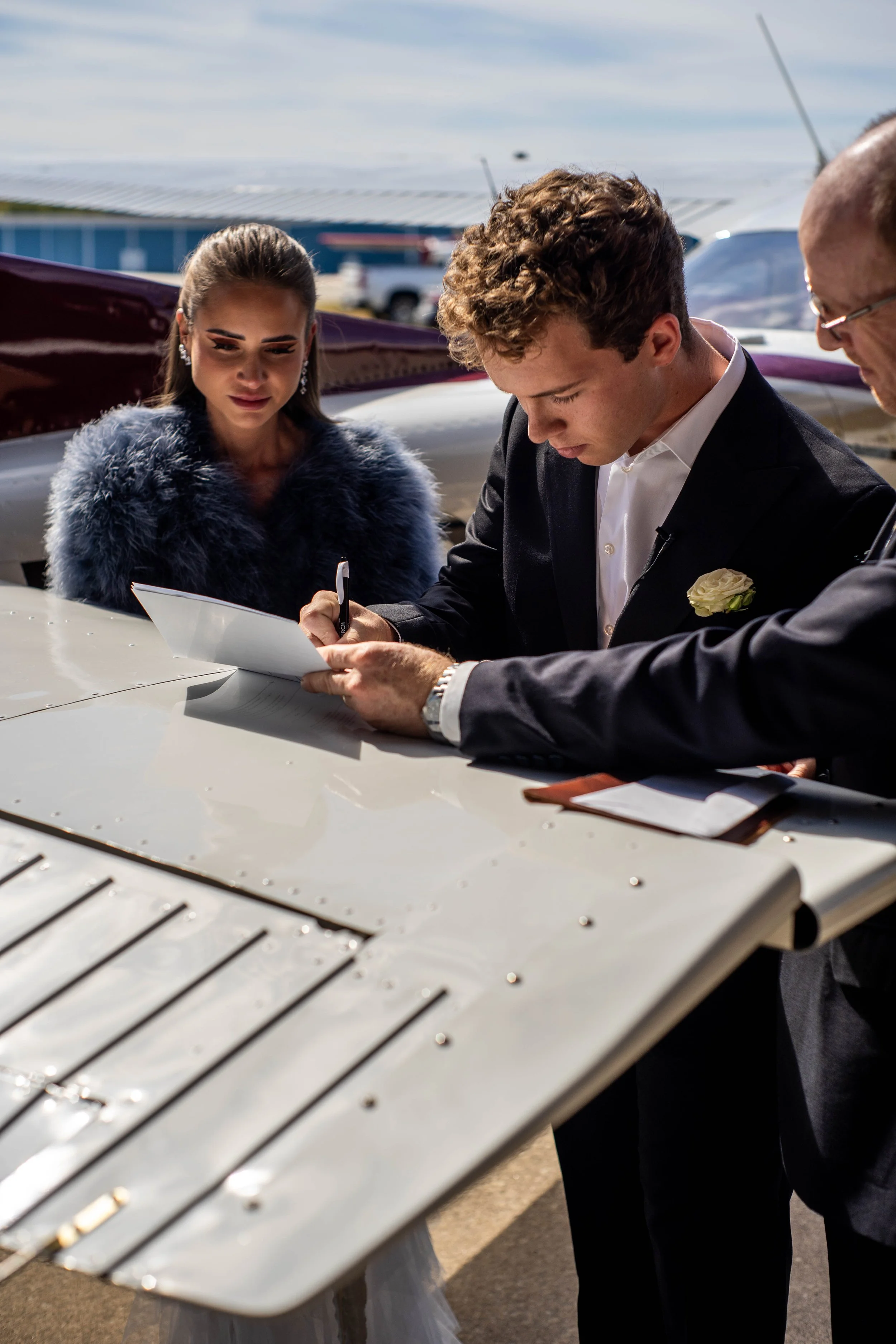A couple at a small aircraft during a wedding, the groom signing a document, the bride observing, with a man assisting, all dressed formally.