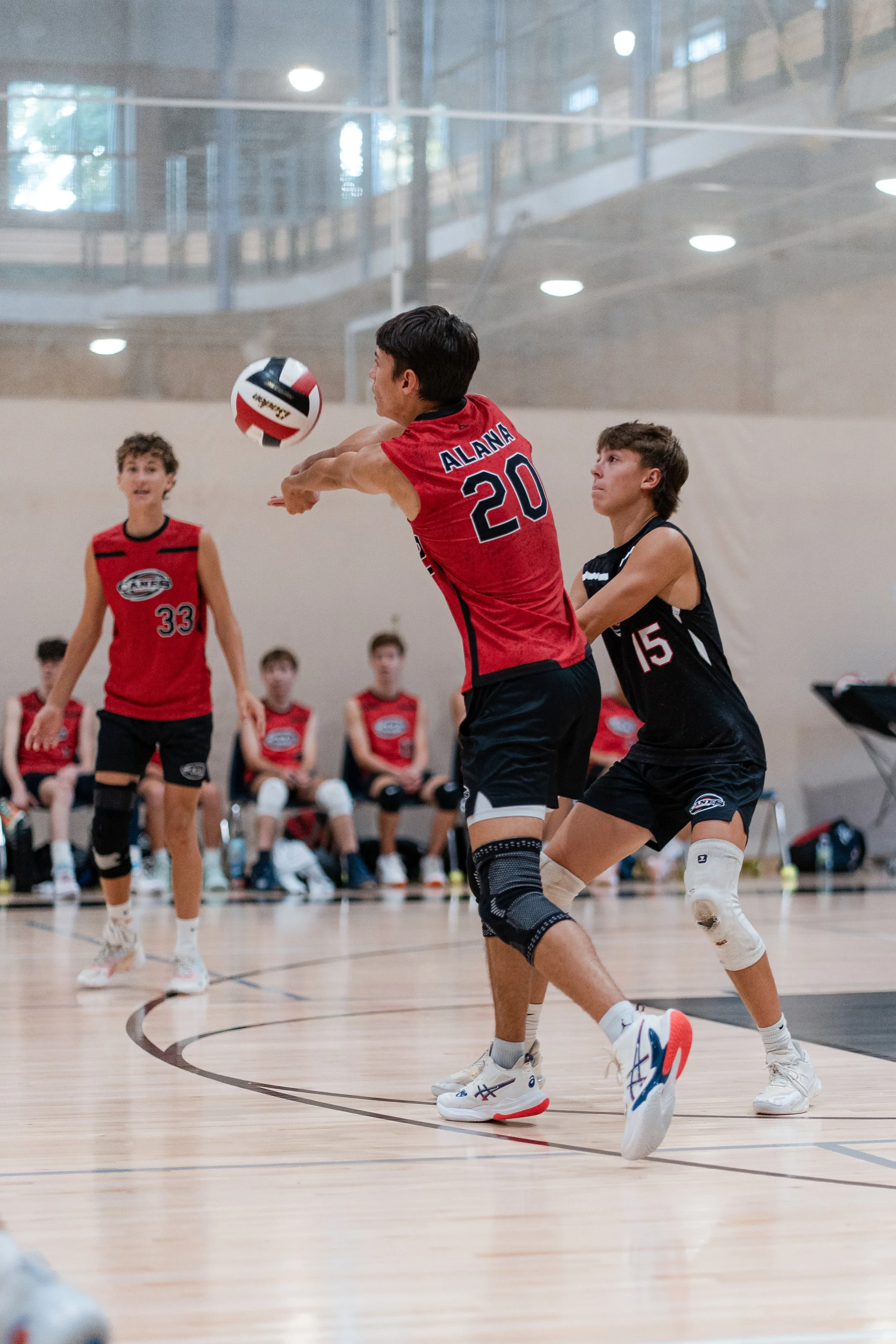 Young volleyball players compete during a game in an indoor gym, with one player wearing a red jersey labeled 'ALANA 20' and two others in black jerseys, one labeled '15,' on the court.